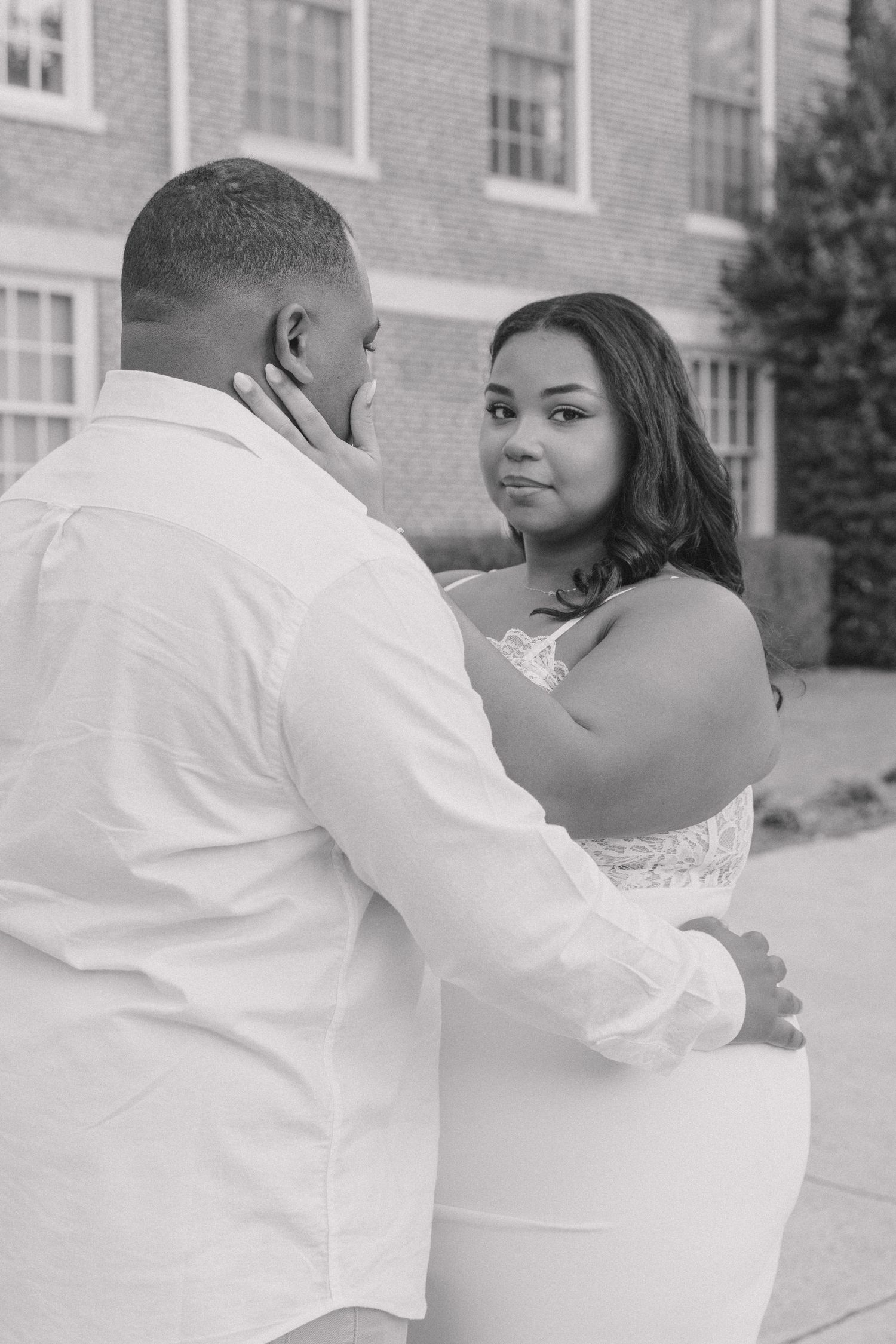 A romantic couple in white outfits embrace and dance together in front of a brick building.