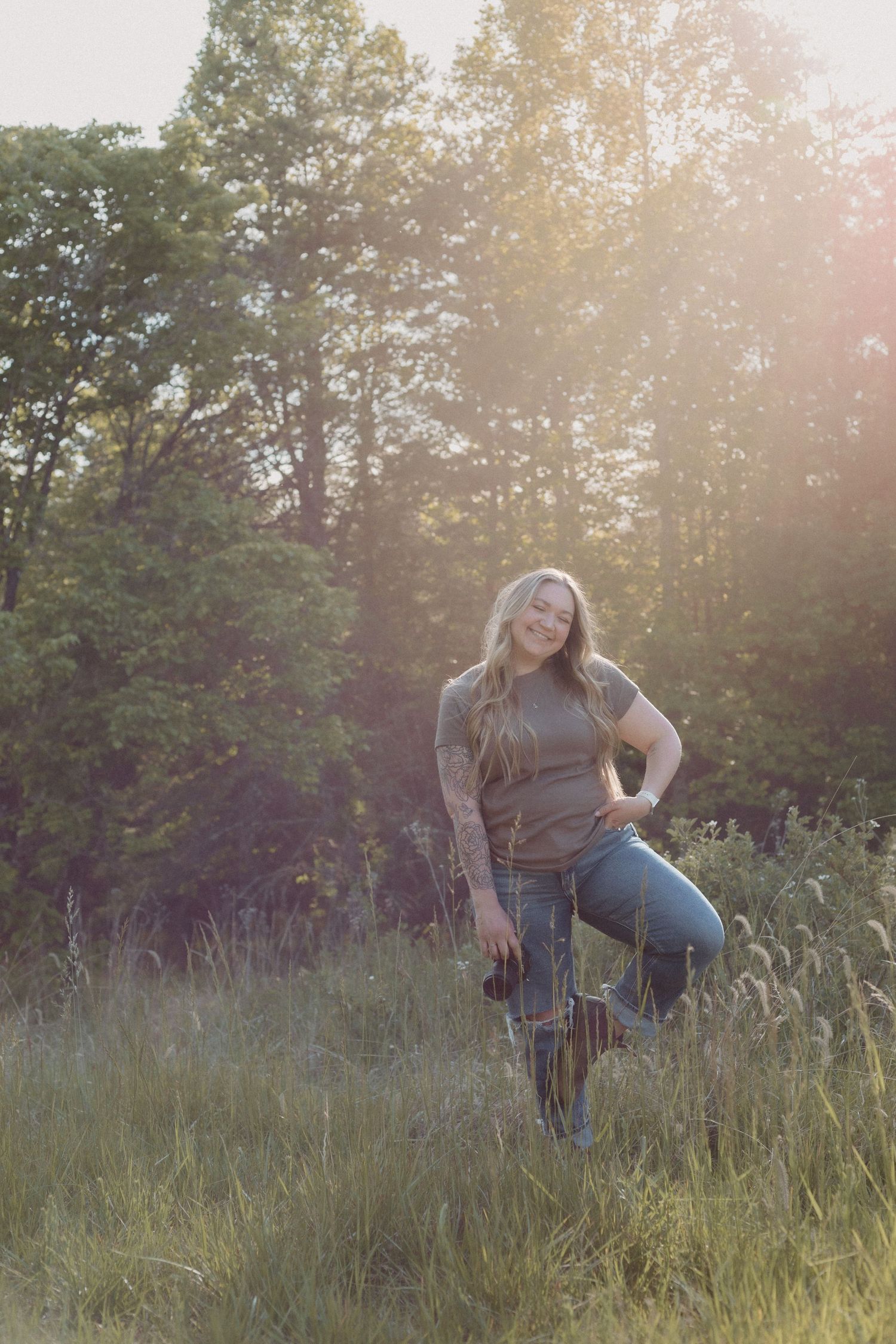 Woman with long blonde hair and tattoos poses in a sunlit field wearing jeans and boots near lush green trees.