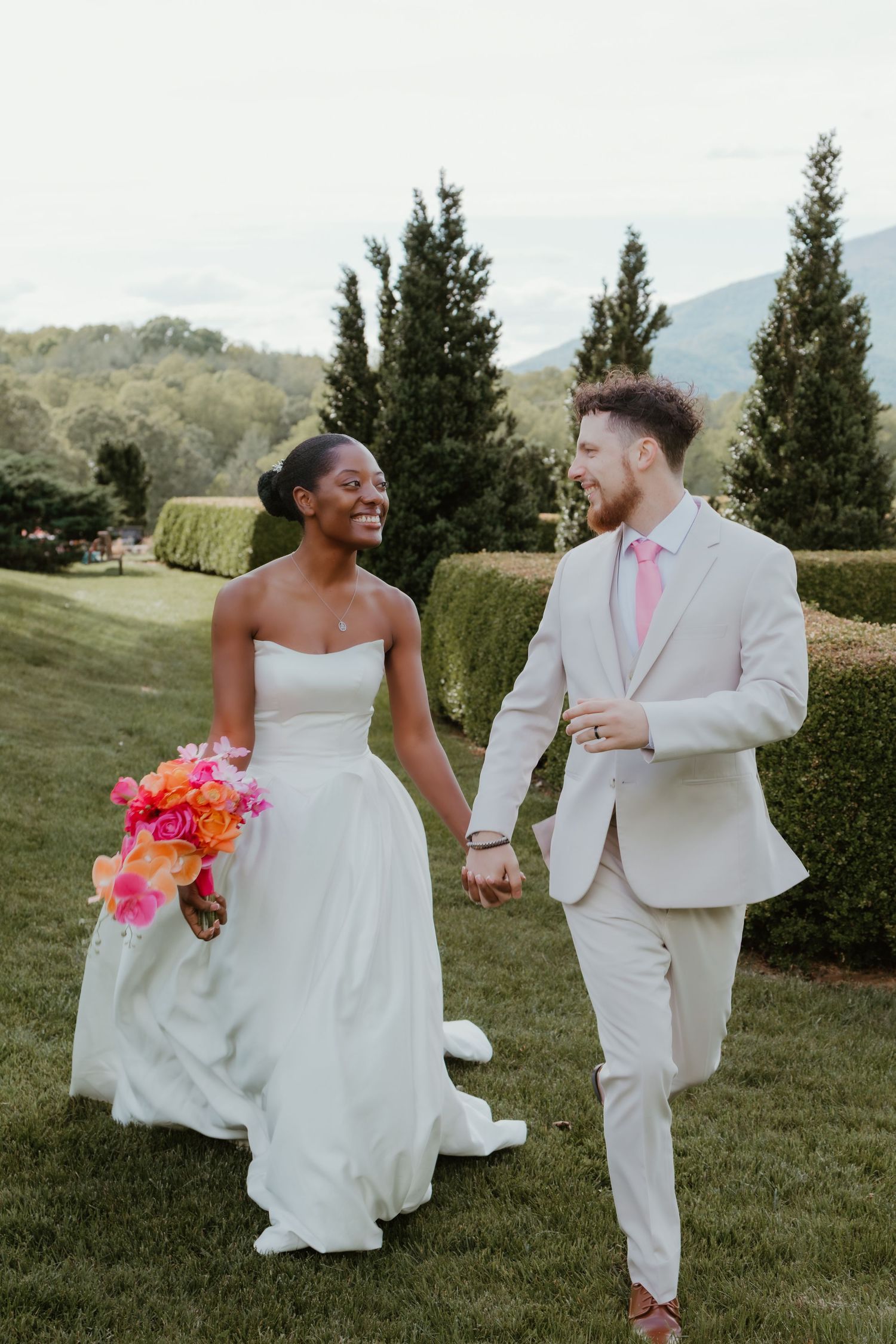 A joyful interracial couple walks hand-in-hand on a lush green lawn, bride holding vibrant pink bouquet in wedding attire.