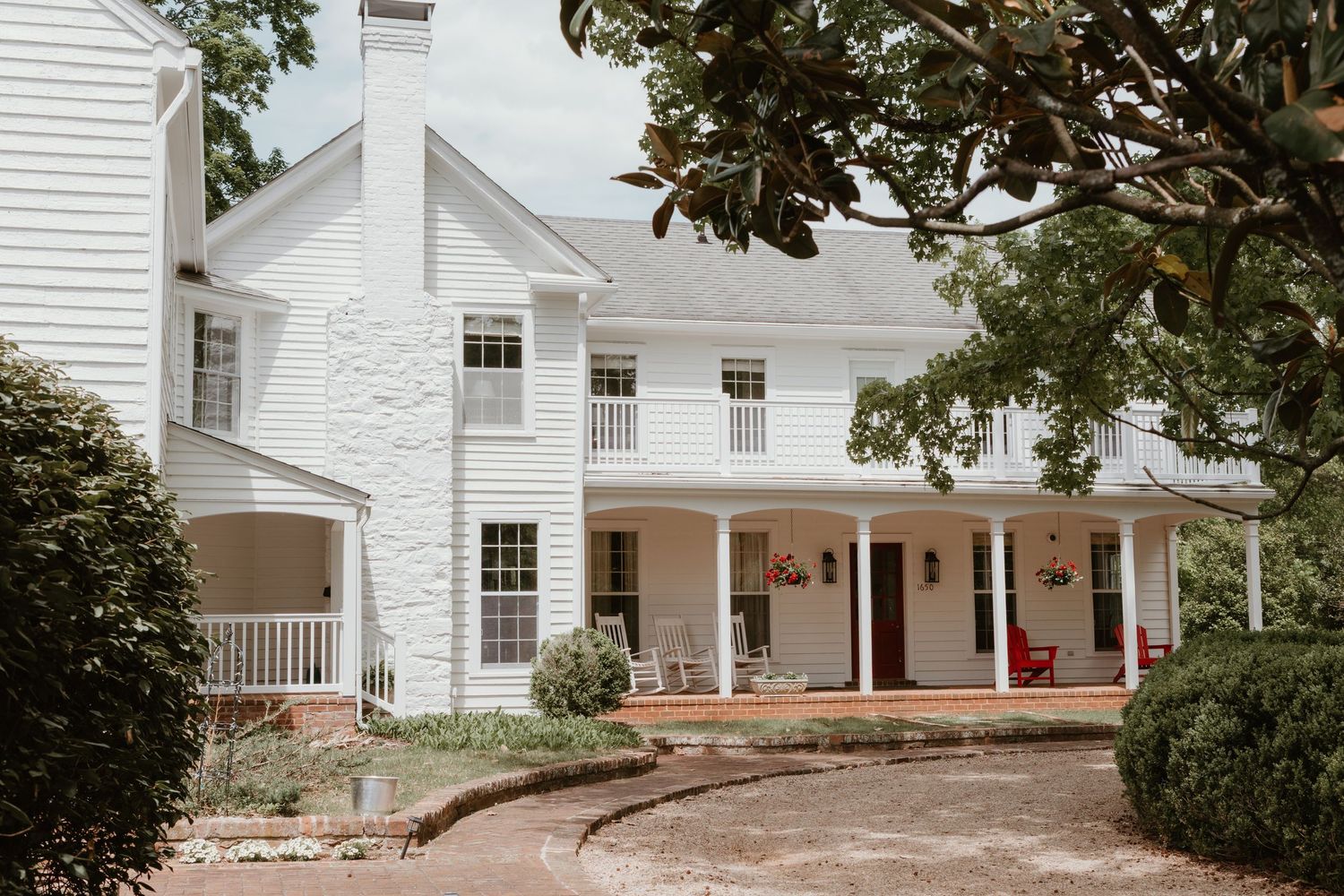 White two-story colonial farmhouse with wrap-around porch, red accents, and lush greenery on a cloudy day.