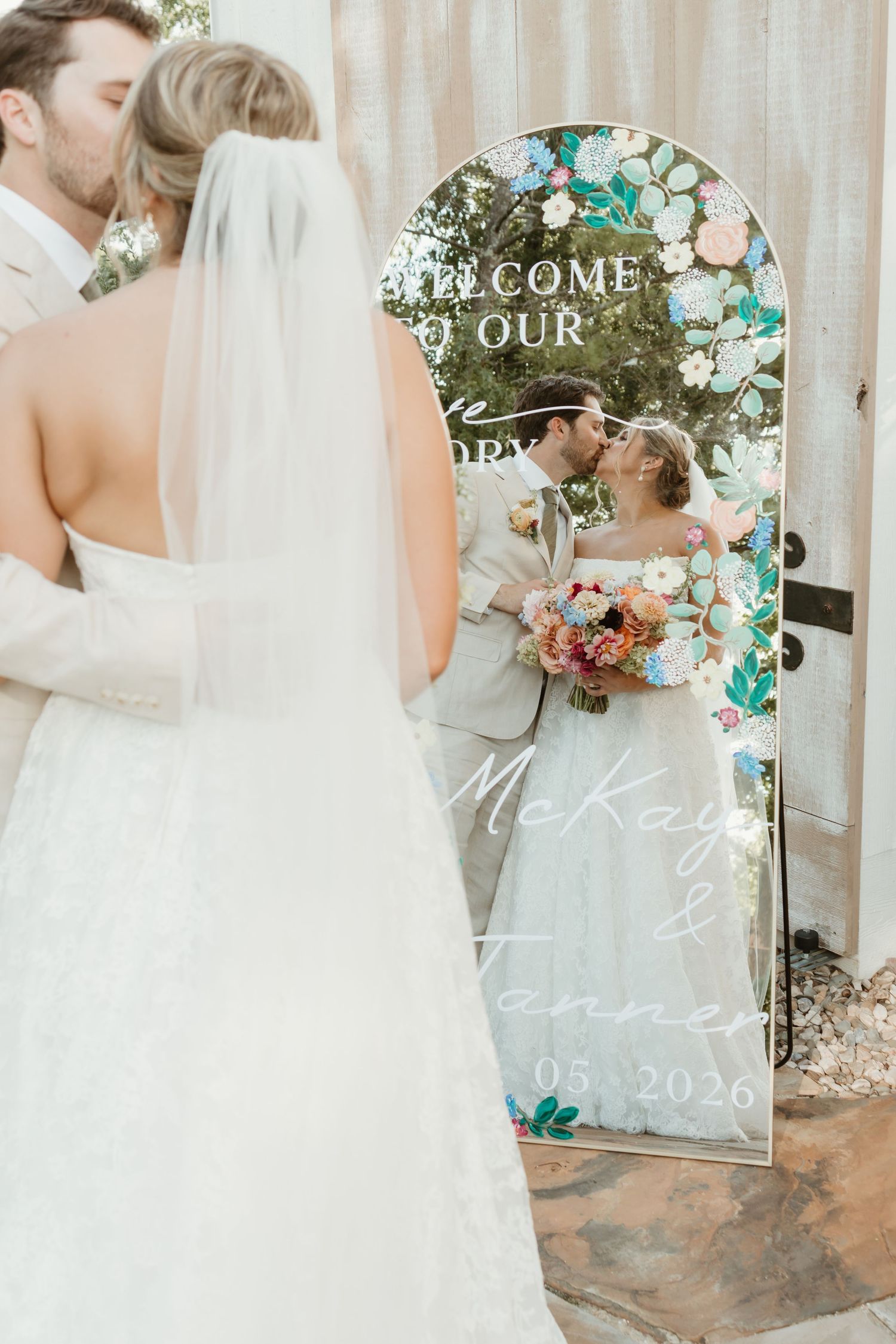 Bride and groom kissing reflected in a floral wedding welcome mirror sign reading McKay and Tanner 05.2026.