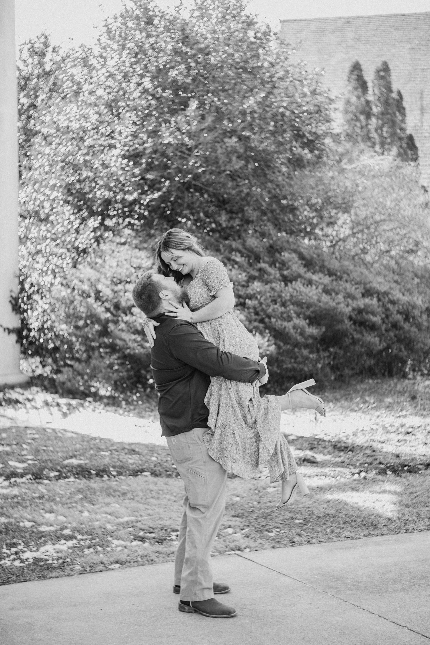 A romantic black and white moment captured outdoors with trees and foliage in the background.