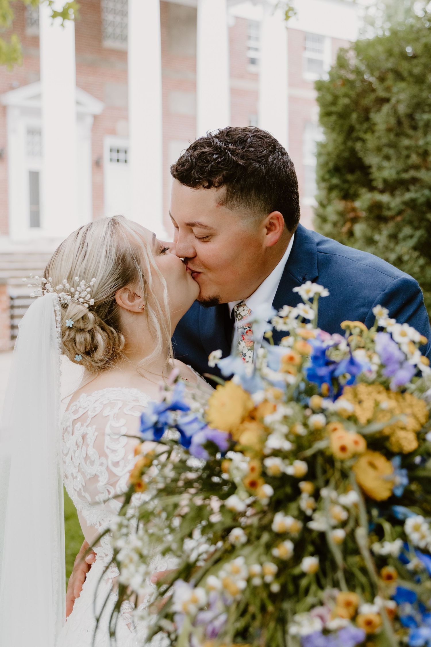 Bride and groom share a kiss holding a colorful wildflower bouquet with blue and yellow blooms outside a brick building.