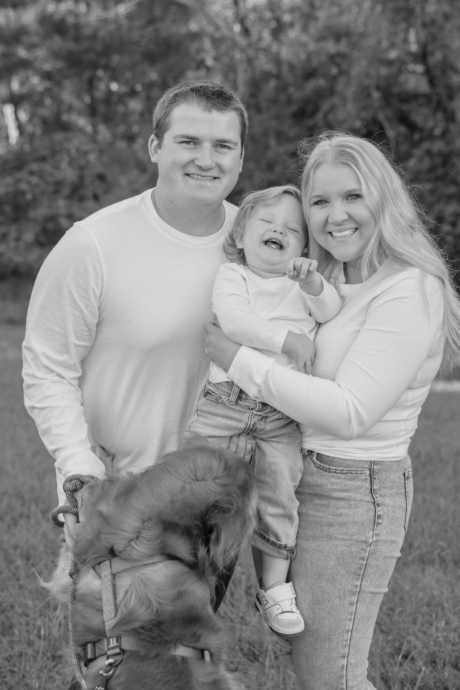 A happy family in white shirts poses together outdoors while their black dog jumps playfully in a series of photos.