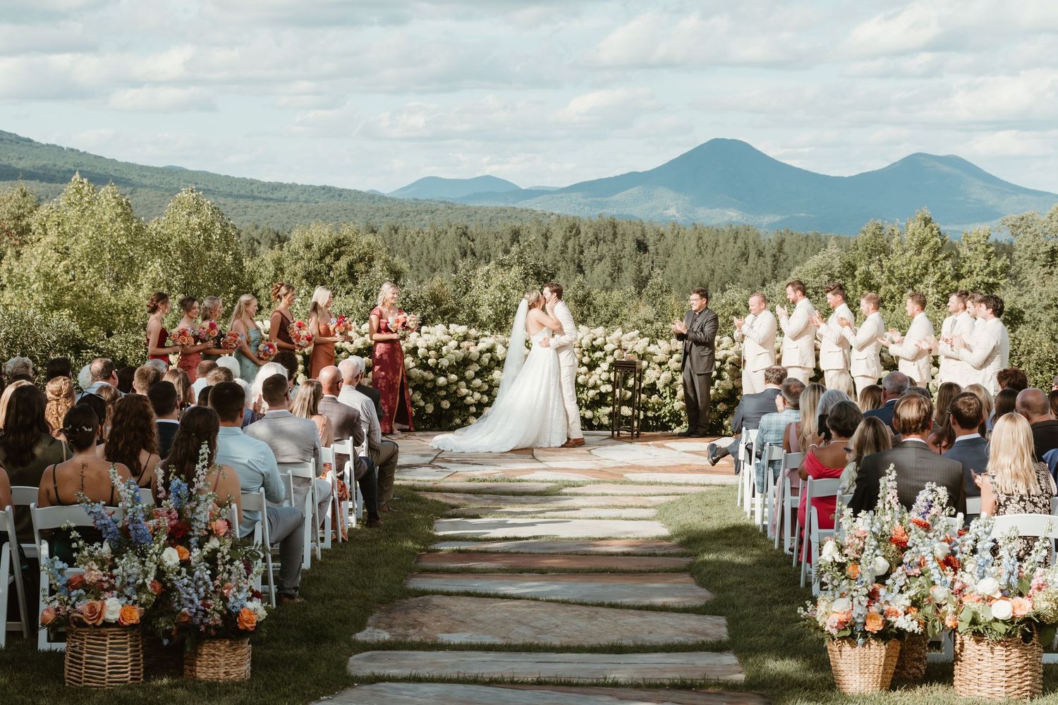 Outdoor wedding ceremony with mountain views, floral aisle decorations, bride and groom at altar, guests seated on both sides.