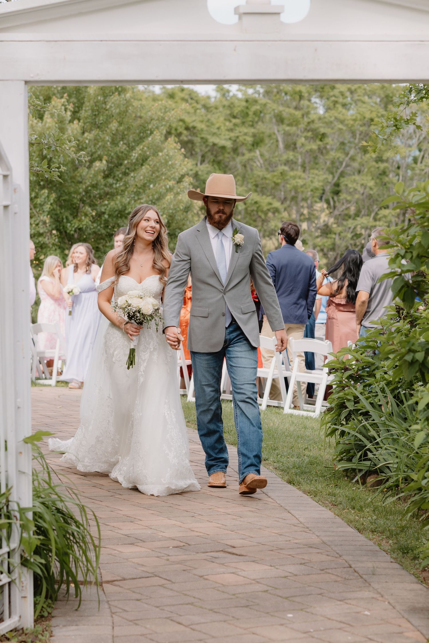 Wedding party members walk down a stone pathway lined with greenery at an outdoor garden ceremony venue.