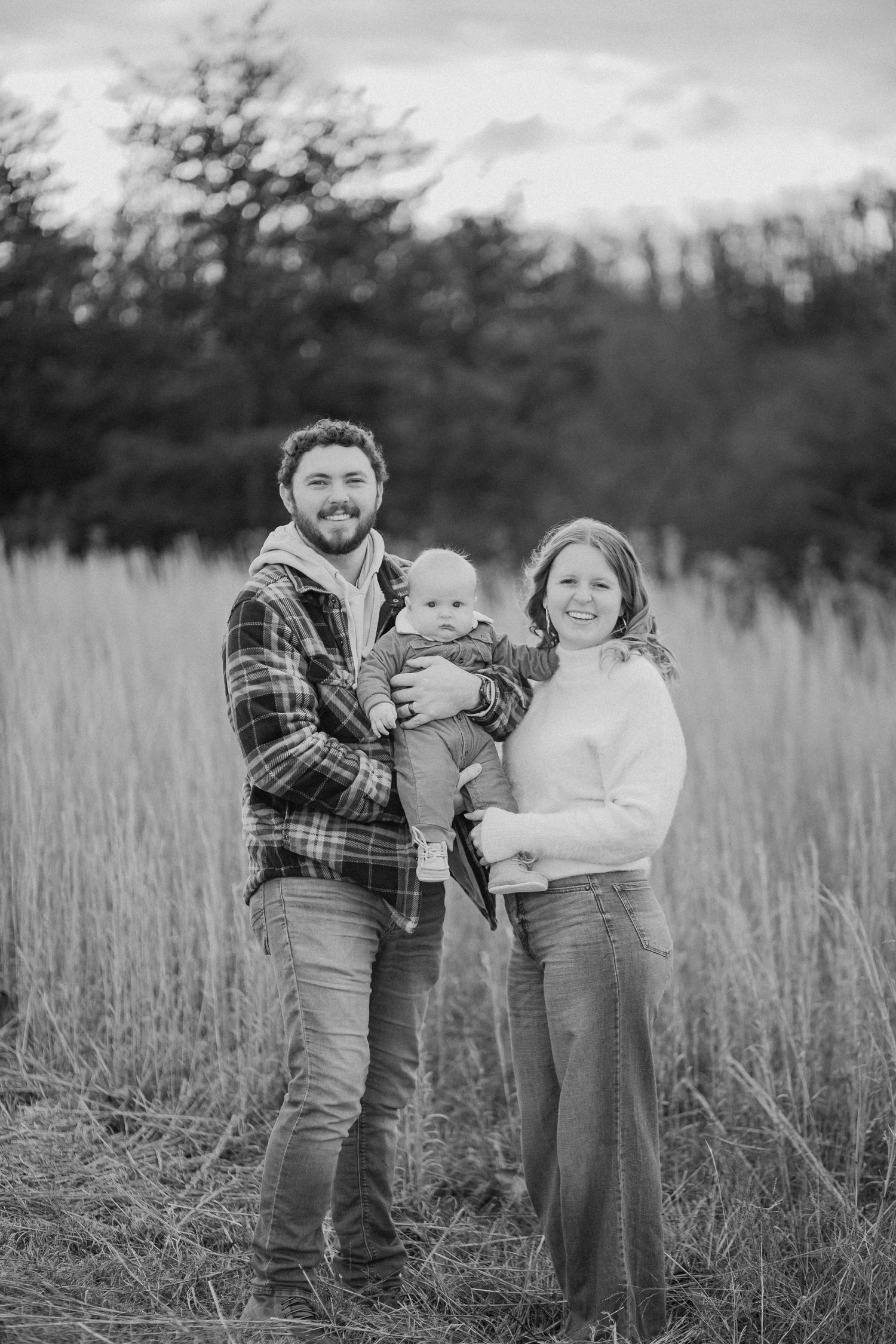 A black and white portrait of a young family standing together in a grassy field surrounded by tall wild grass.