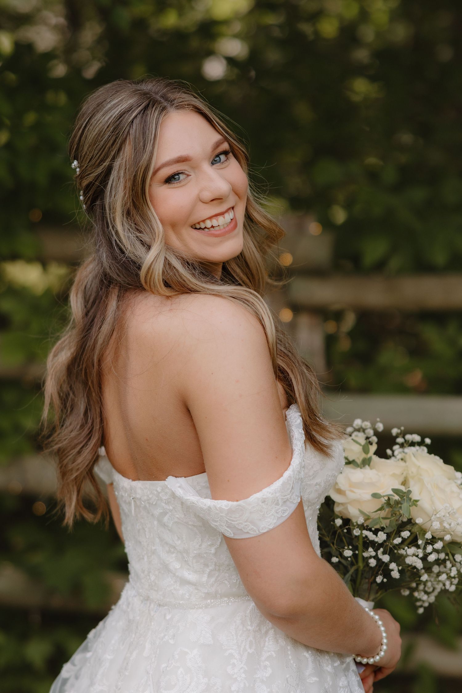 A bride in a strapless white lace wedding dress poses outdoors with baby's breath flowers in a natural garden setting.