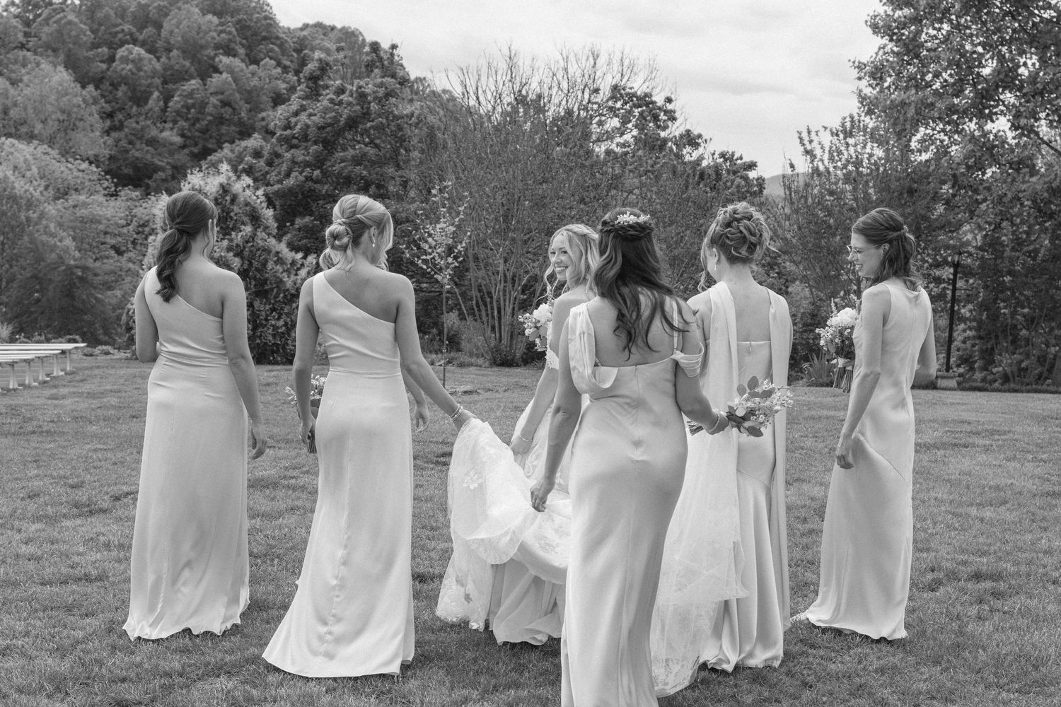 Black and white photo of a bride and bridesmaids in white gowns standing together on a green lawn outdoors.