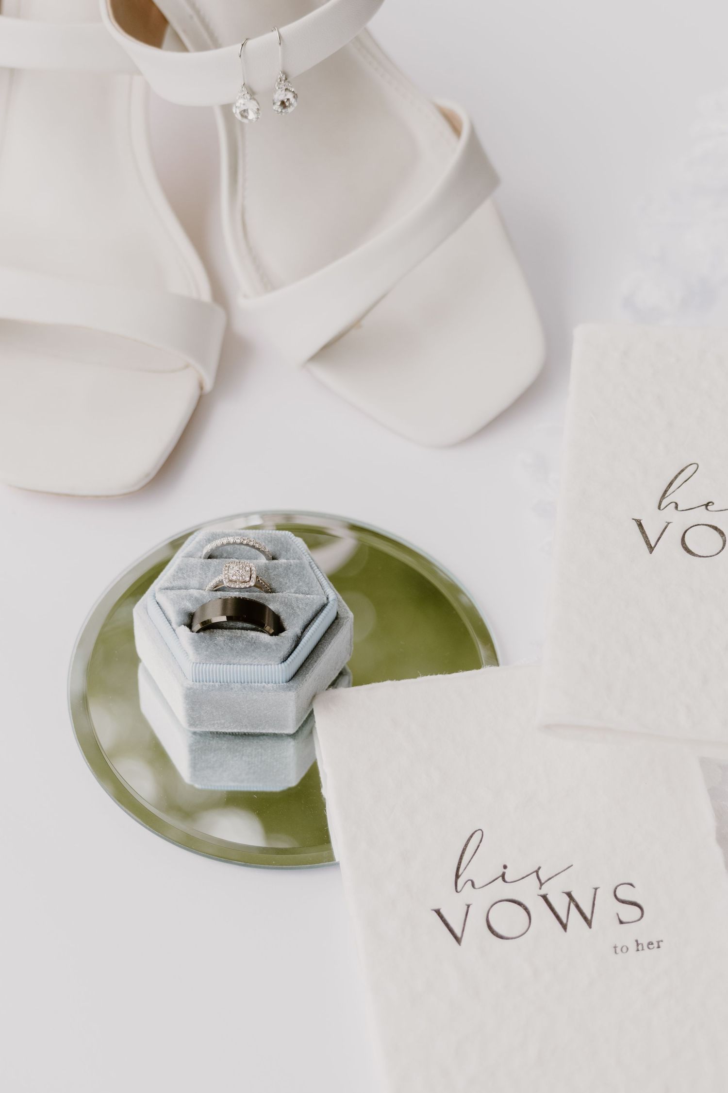 White bridal heels, diamond engagement ring in velvet box, and his vows booklets styled on elegant white wedding flat lay.