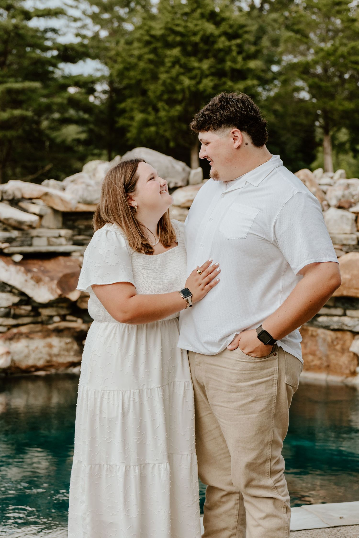 A couple in white and beige outfits smiling at each other near a stone waterfall and pool outdoors.