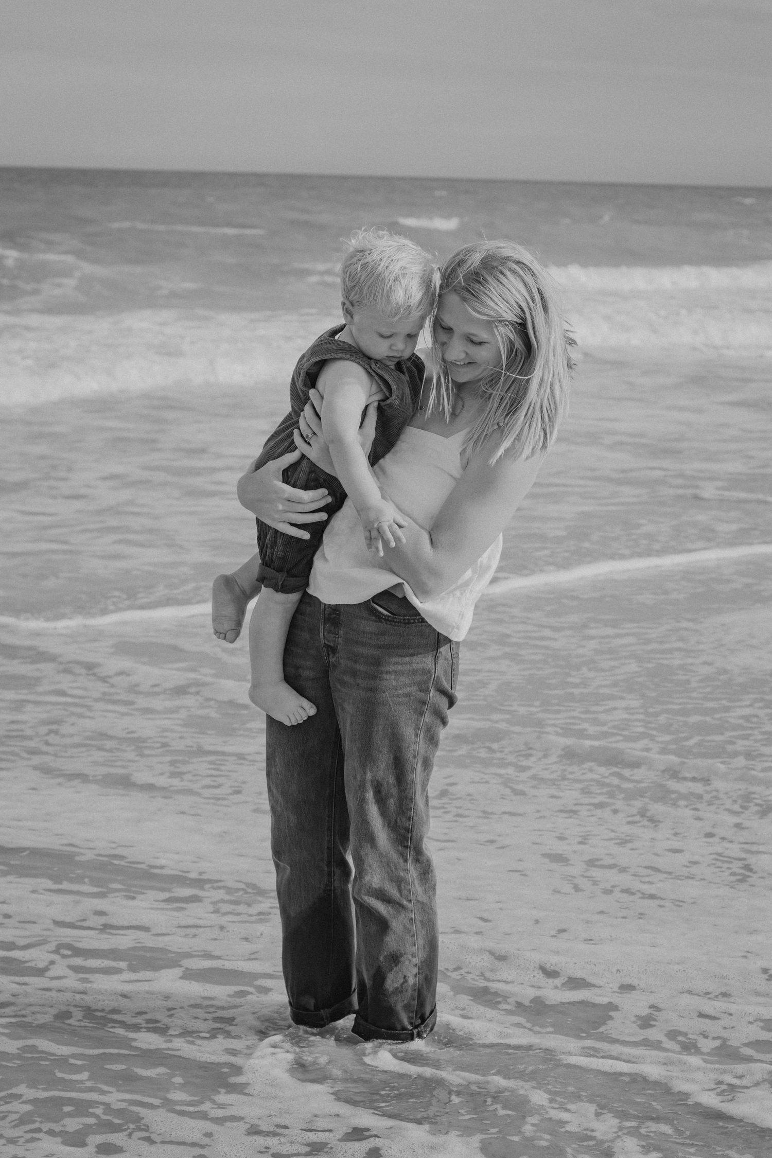 A tender black and white beach moment as parent and child embrace in the surf while waves roll in behind them.