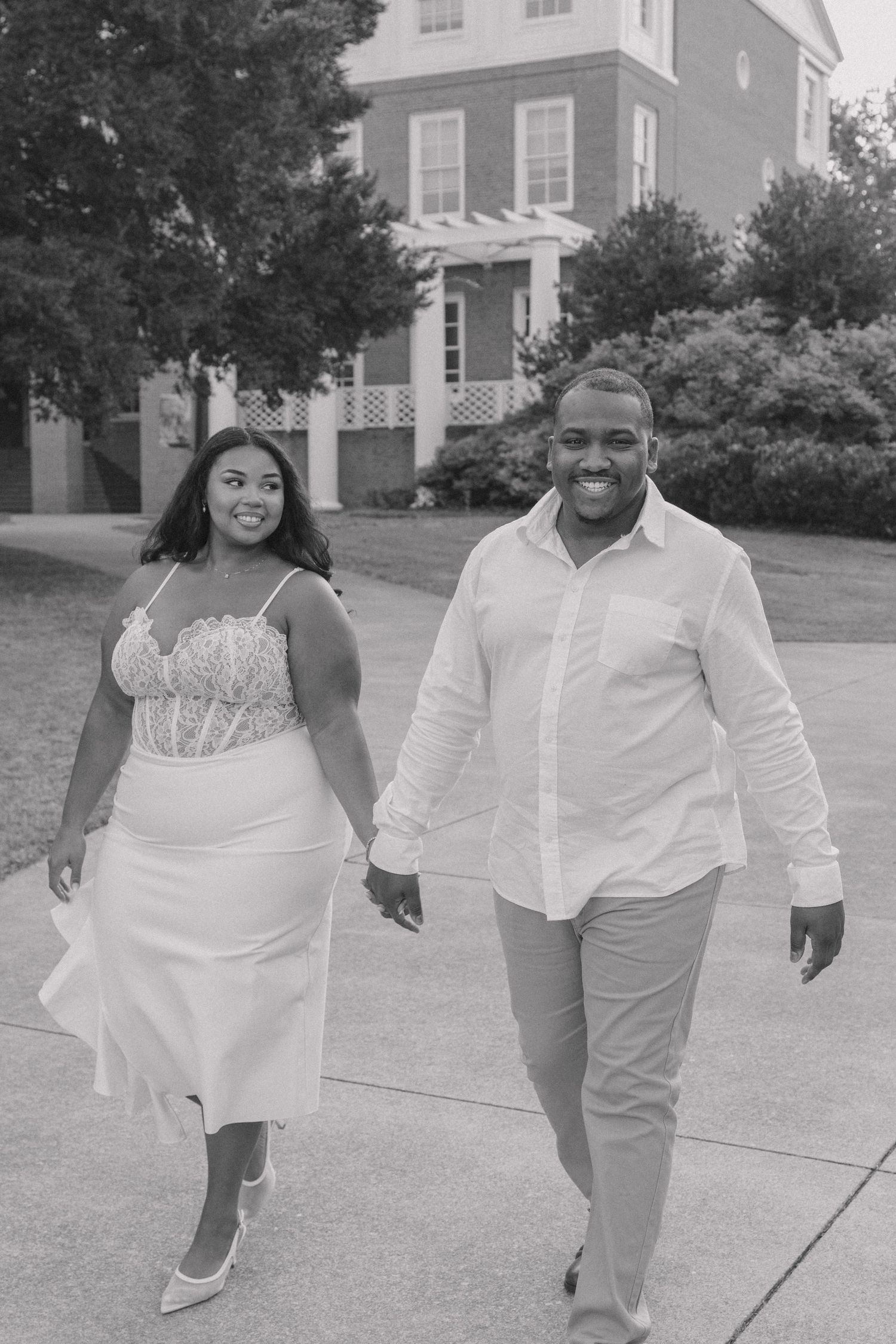 A couple in white outfits walking and dancing playfully together on a paved walkway in black and white.