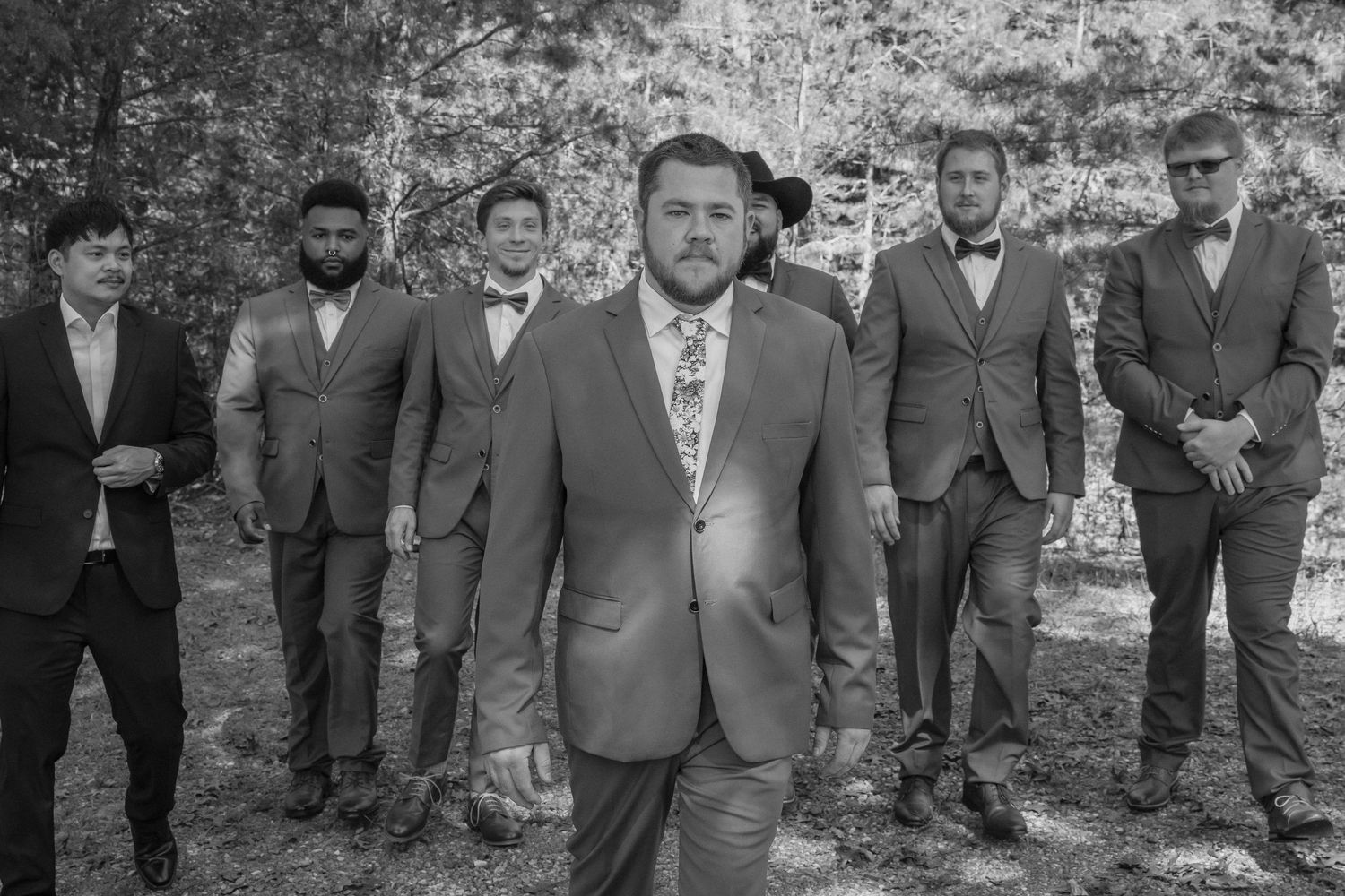 A group of groomsmen in suits pose outdoors in a black and white photo with a natural wooded backdrop.