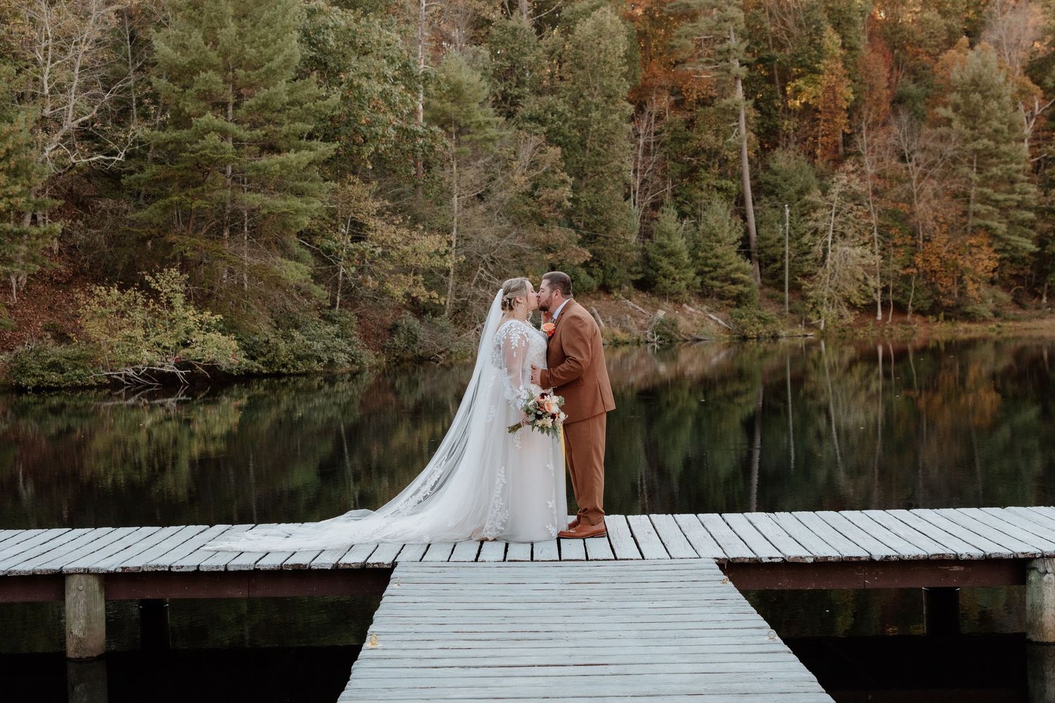 A couple stands on a white wooden dock overlooking a serene lake surrounded by autumn trees.