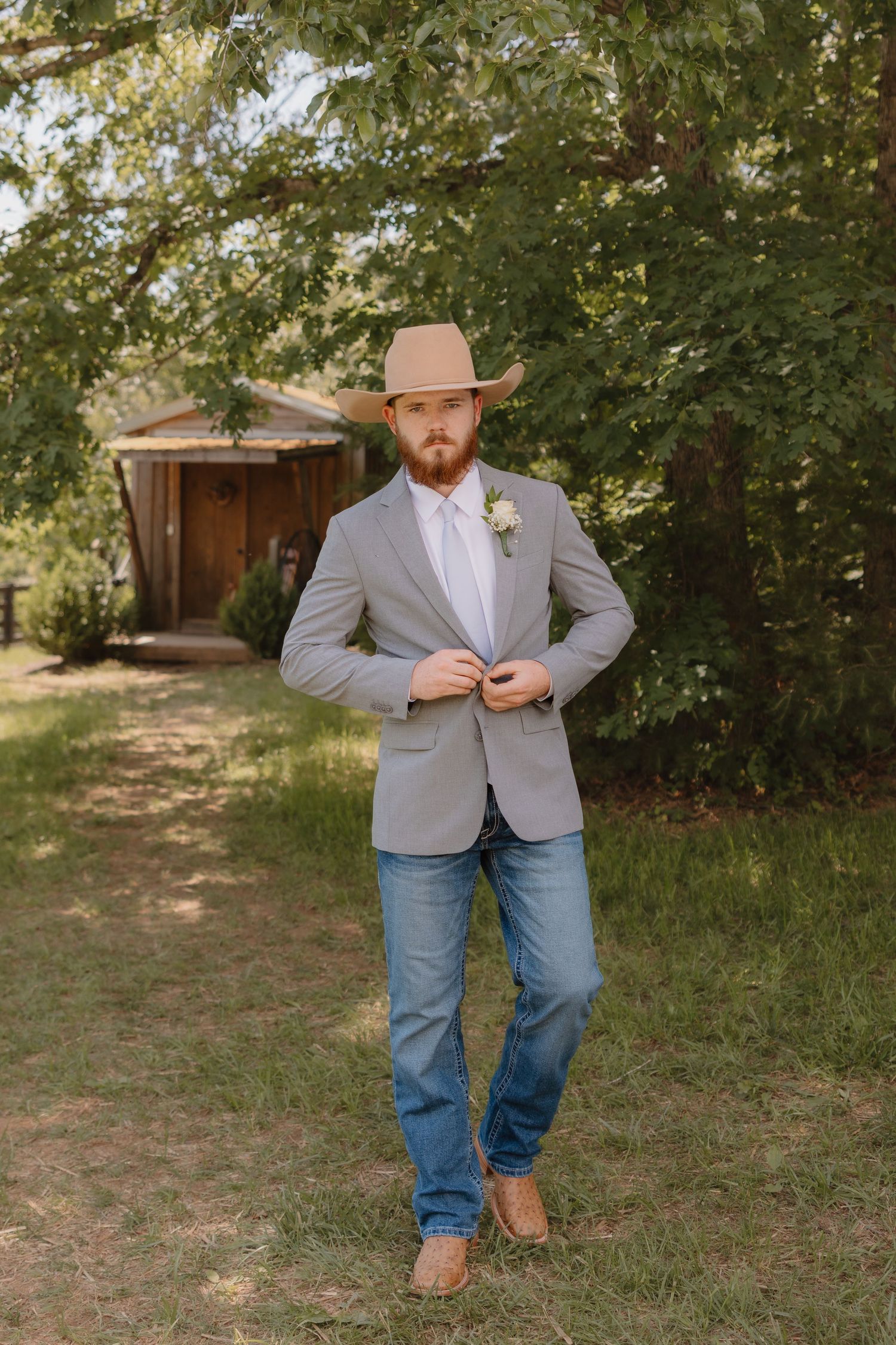 A groom in a gray blazer, jeans, and cowboy boots poses casually in an outdoor garden setting.