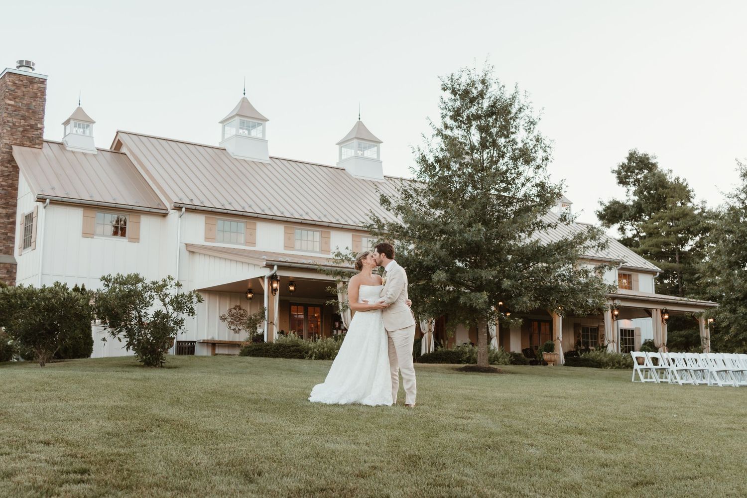 Bride and groom share a romantic kiss on lush green lawn in front of elegant white barn wedding venue.