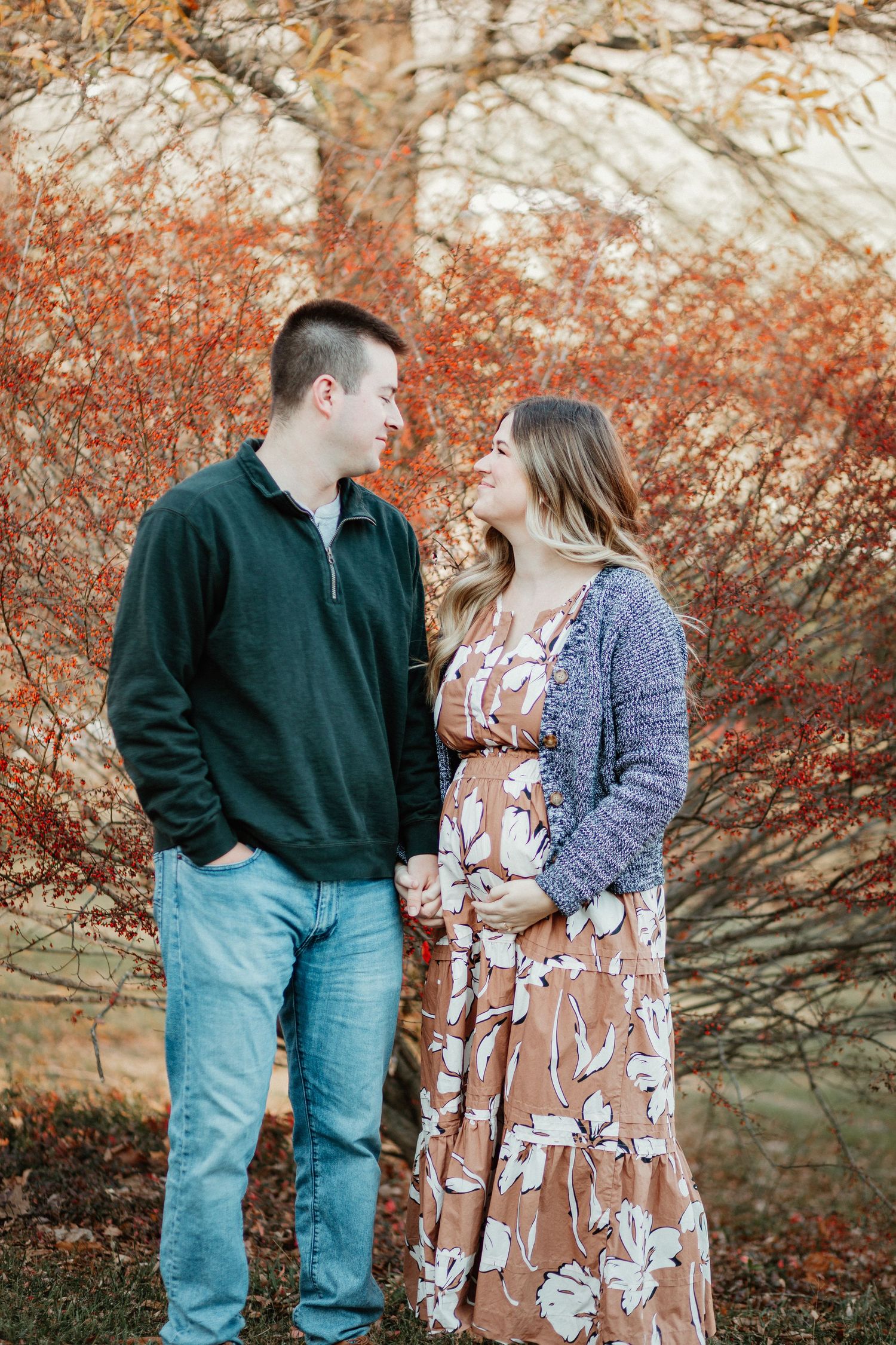 A couple in casual fall attire stands together in front of autumn foliage with orange and red colors.