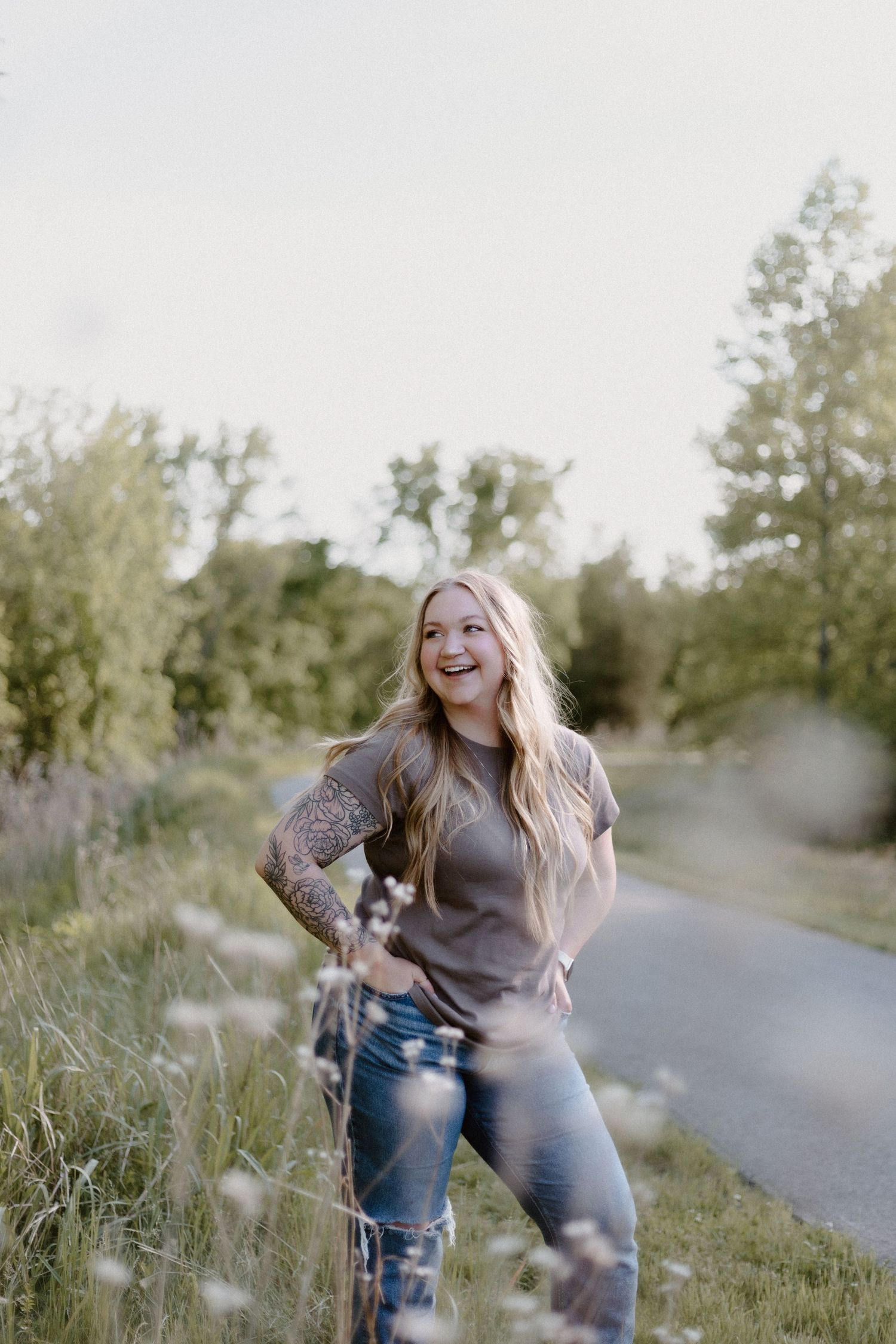 Smiling blonde woman with tattoos in jeans and gray tee standing on a rural path surrounded by wildflowers and trees.