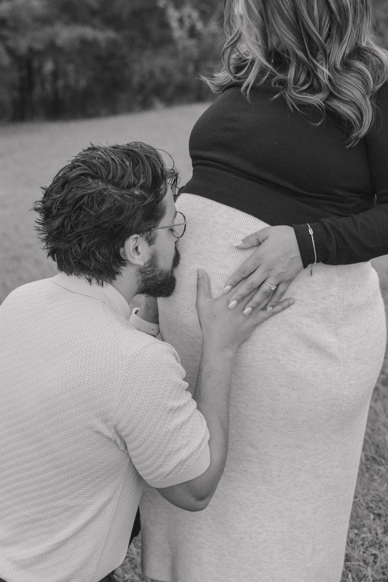 A tender black and white maternity photo shows an expectant couple sharing an intimate moment outdoors.