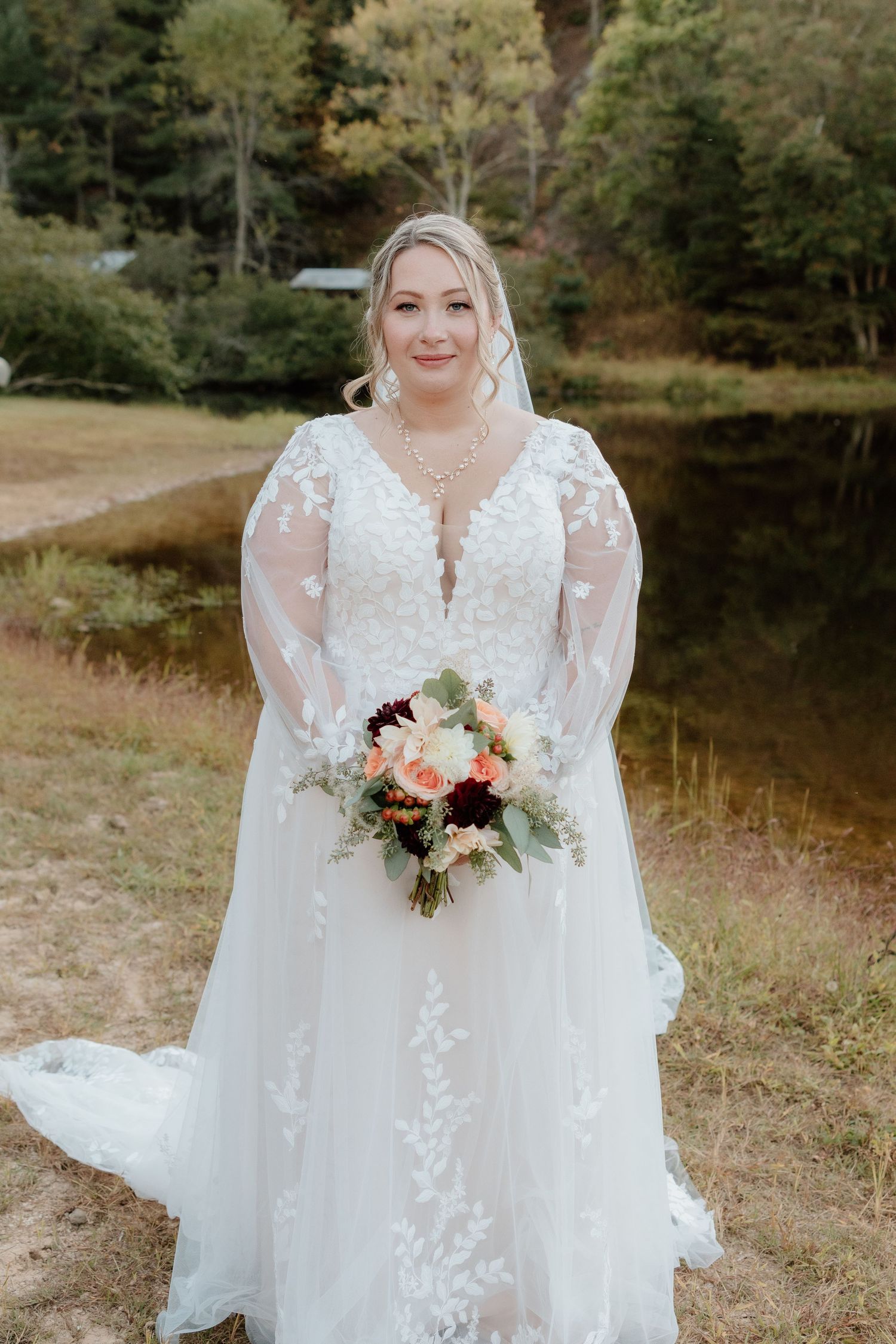 A bride in a flowing white lace gown with sheer sleeves holds a romantic bouquet of pink and white flowers by a lake.