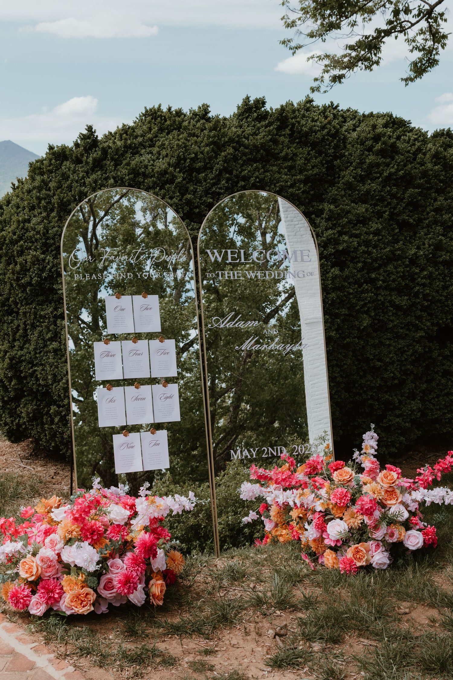 Two arched gold mirror wedding signs with seating chart and welcome message, adorned with vibrant pink and coral floral arrangements.