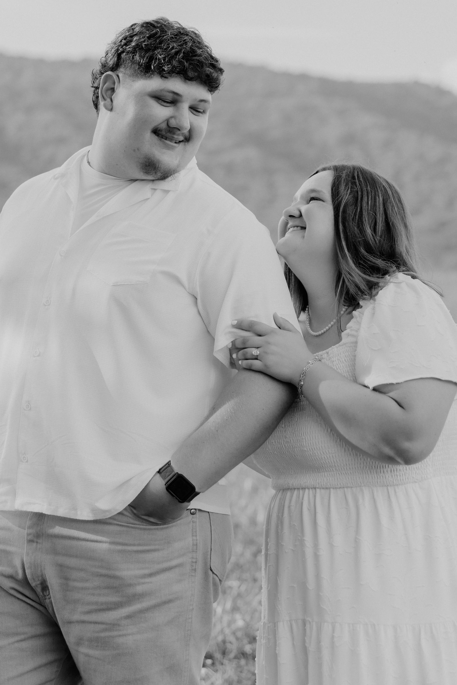 Black and white photo of a happy couple holding hands outdoors with mountains in the background.
