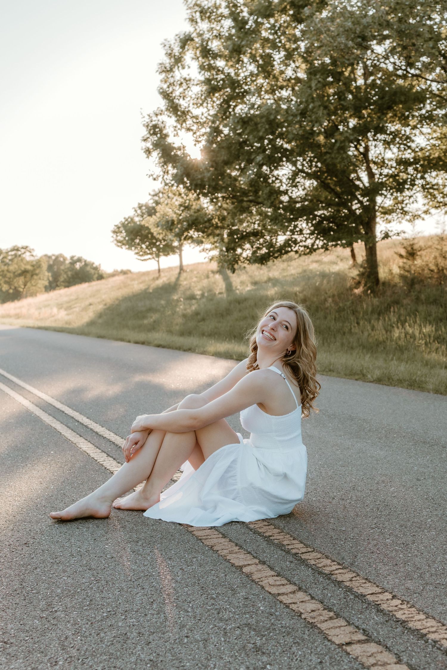 Young woman in white dress sitting barefoot on a quiet road, smiling, with golden hour sunlight and green trees behind her.