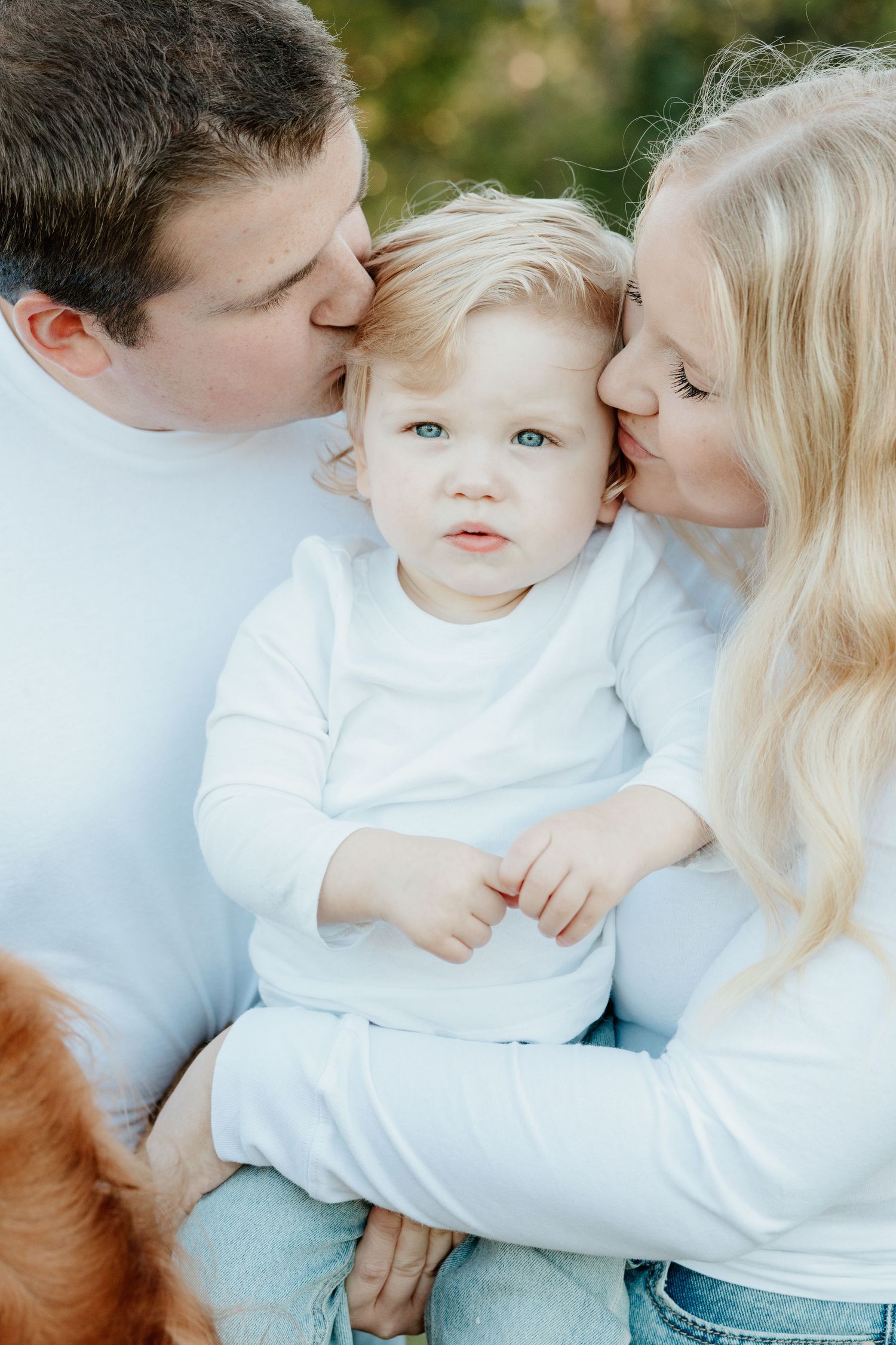 A family shares tender kisses and embraces while wearing coordinating white and light blue outfits in an outdoor setting.