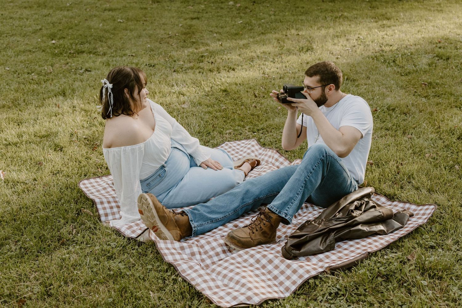 Couple sitting on a plaid picnic blanket in a park, man photographing woman with a vintage camera on green grass.