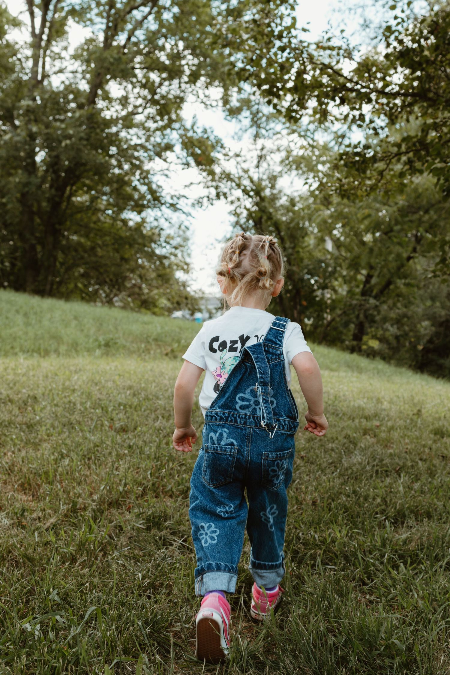 Toddler girl in denim overalls and white tee walks across green grass toward trees in a park.