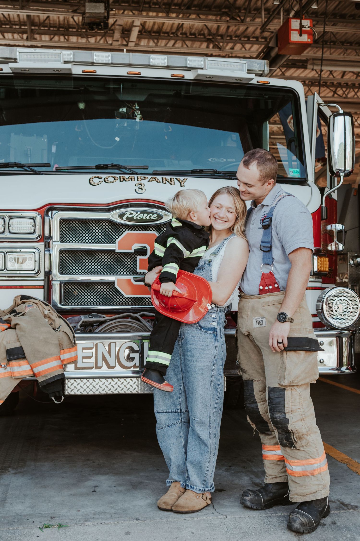 A family poses in front of a fire engine, with a firefighter kissing his wife while their child wears a toy helmet.
