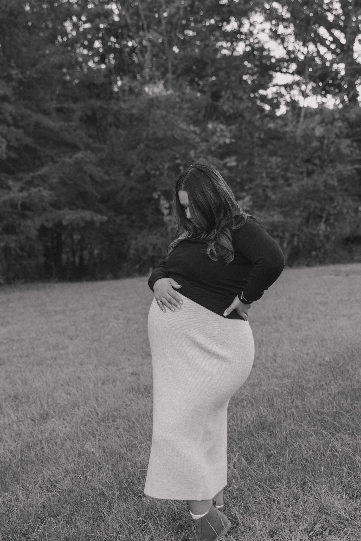 A person in a black top and white skirt poses outdoors in a grassy area with trees in the background.