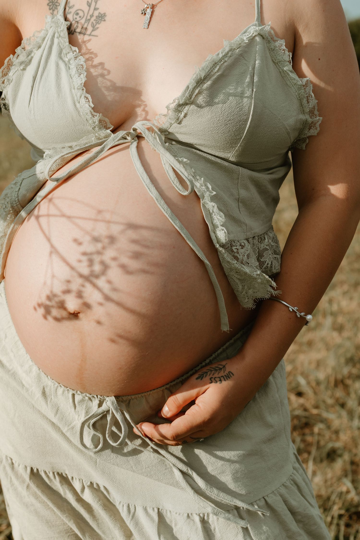 Pregnant woman in sage green lace maternity bralette and skirt, cradling bare baby bump outdoors with floral shadow.