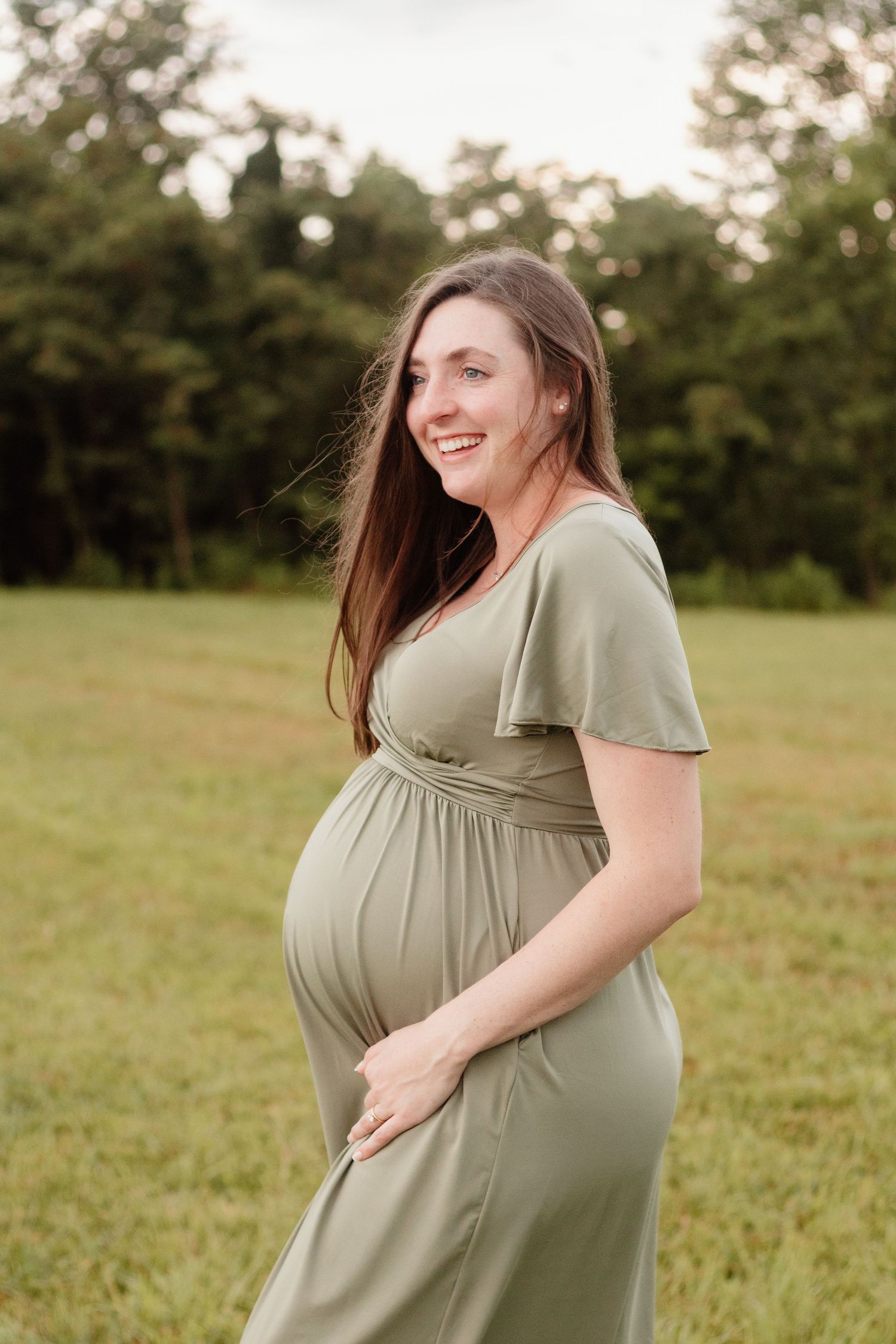 Smiling pregnant woman in olive green maternity dress standing in a grassy field during outdoor maternity photo session.