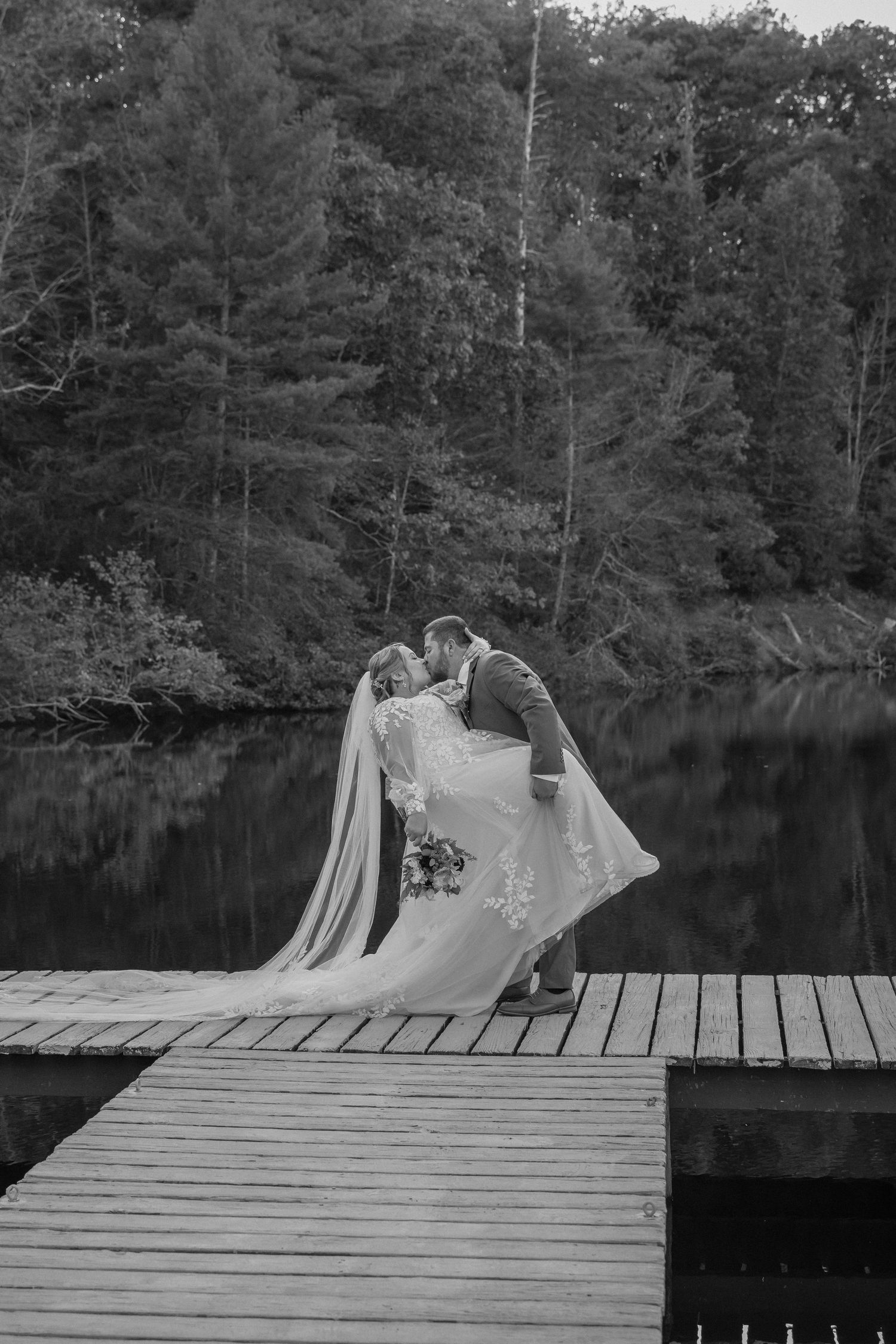 A romantic wedding moment captured in black and white on a wooden dock overlooking a serene lake surrounded by trees.