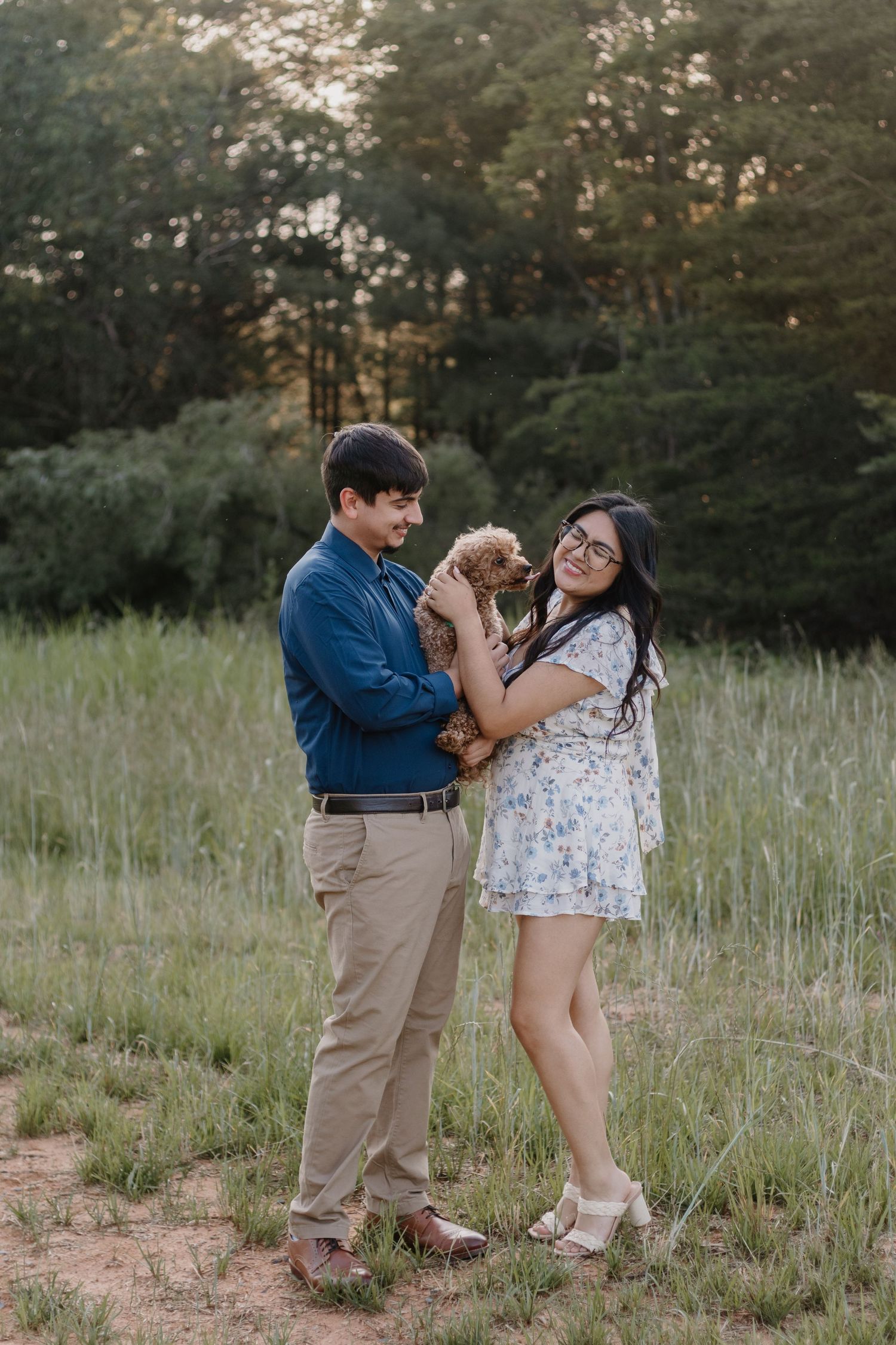 Couple smiling and holding a small brown puppy in a green meadow with trees in the background.