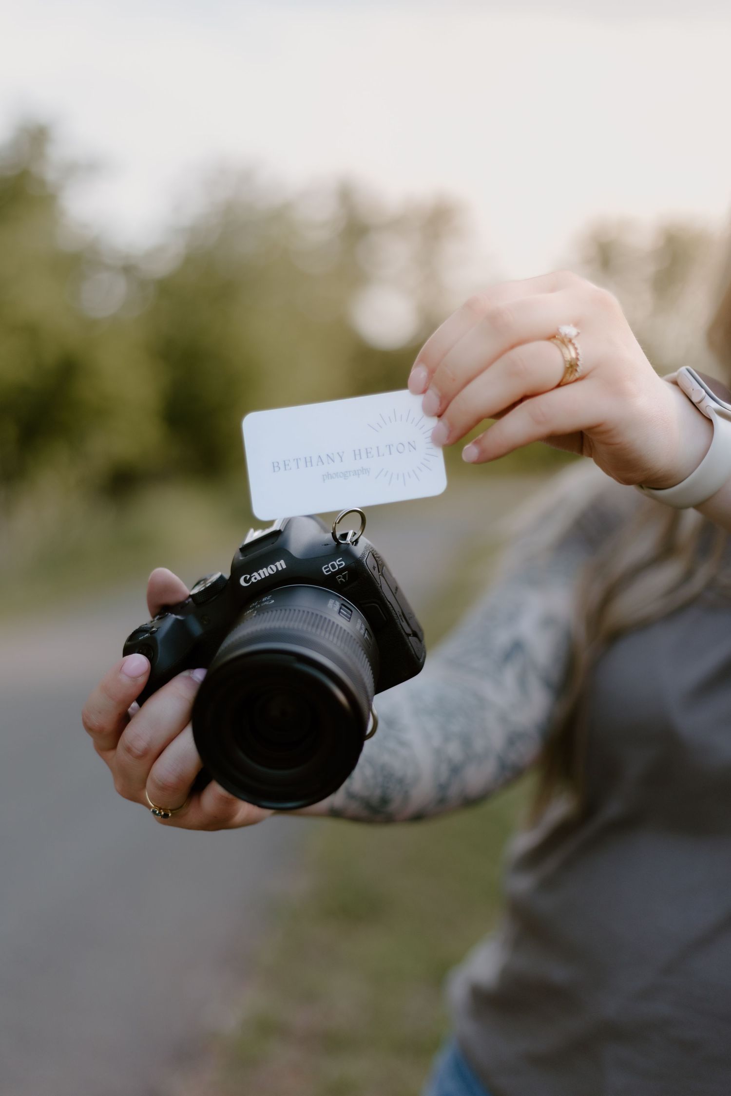 Person holding a Canon EOS camera and a business card outdoors, with a tattooed arm and gold ring visible.
