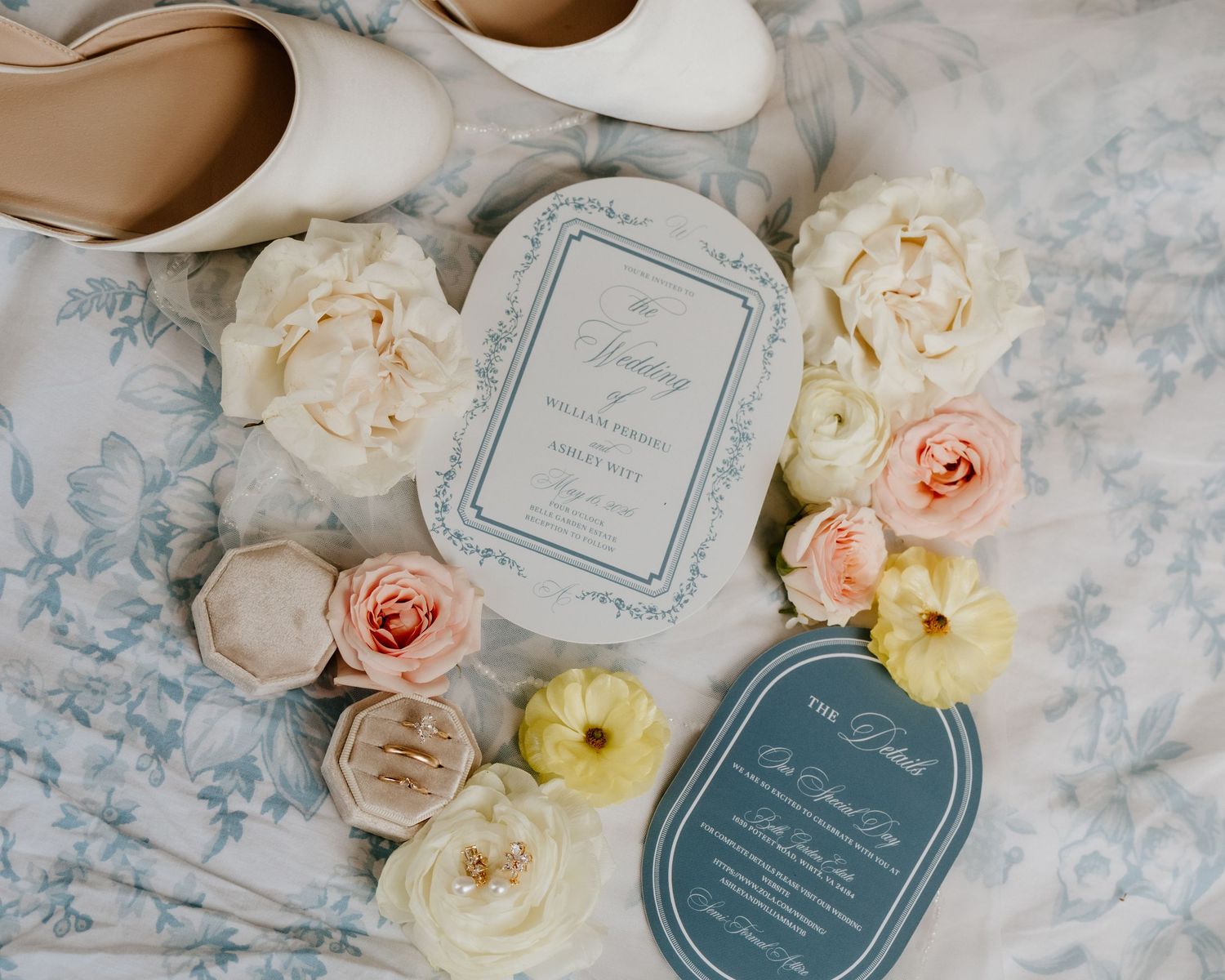 Elegant wedding flat lay with blush roses, white heels, and blue stationery on floral fabric.