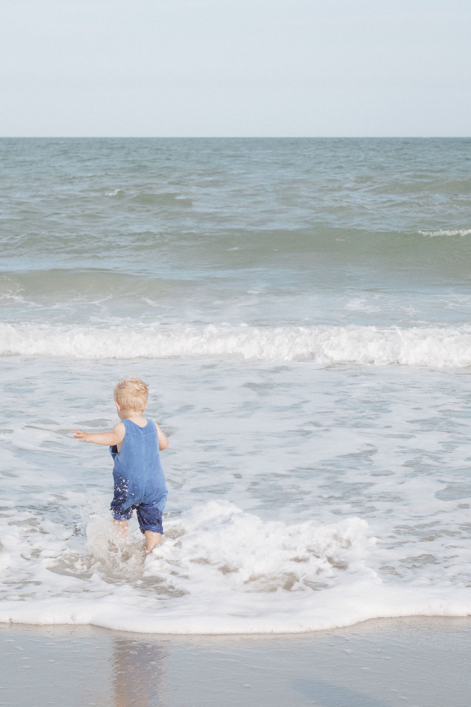 A small child in blue clothing splashes and plays in the ocean waves at the beach.