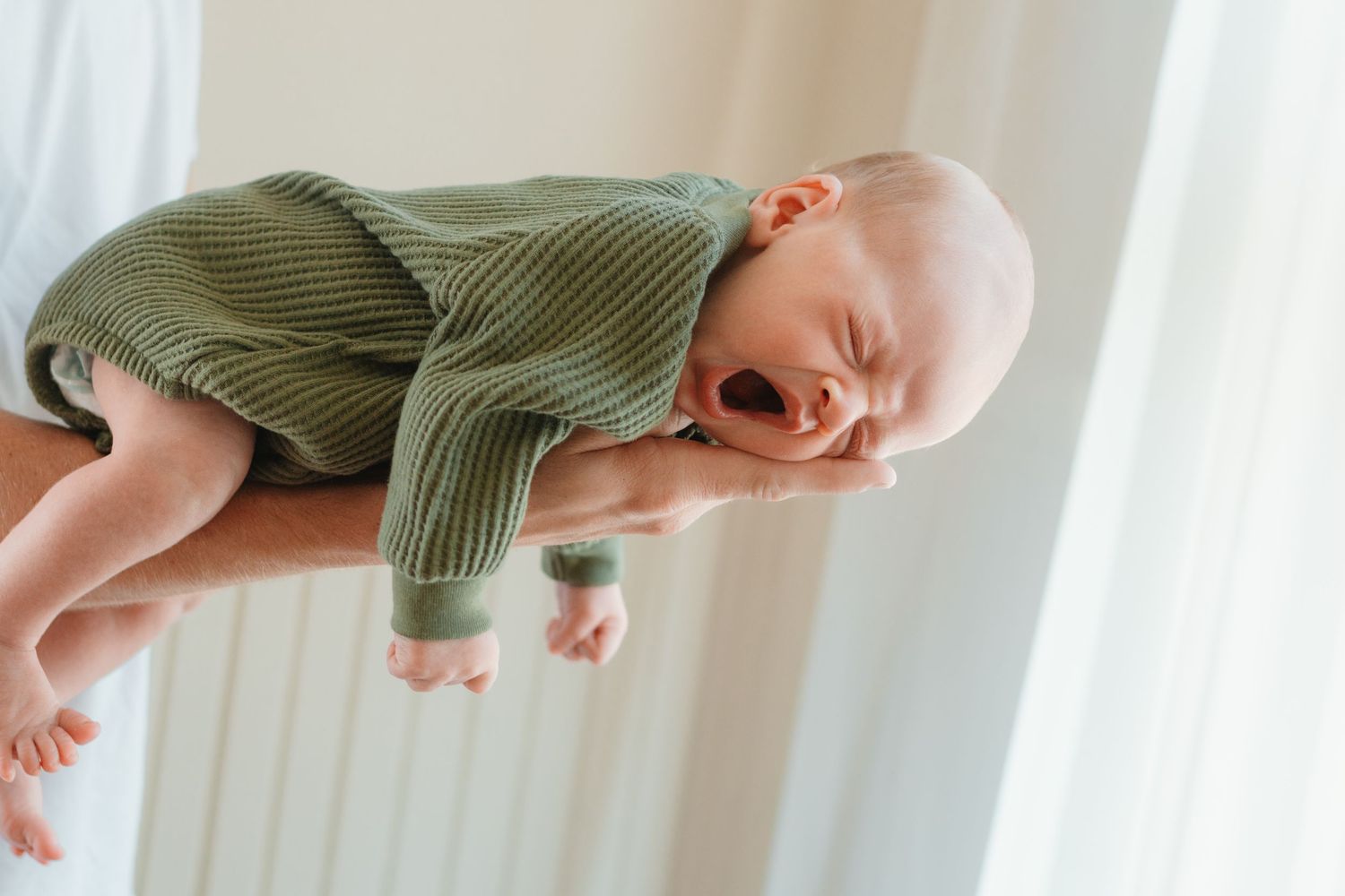 A crying newborn baby in a green ribbed romper being held horizontally on an adult's arm near a bright window.