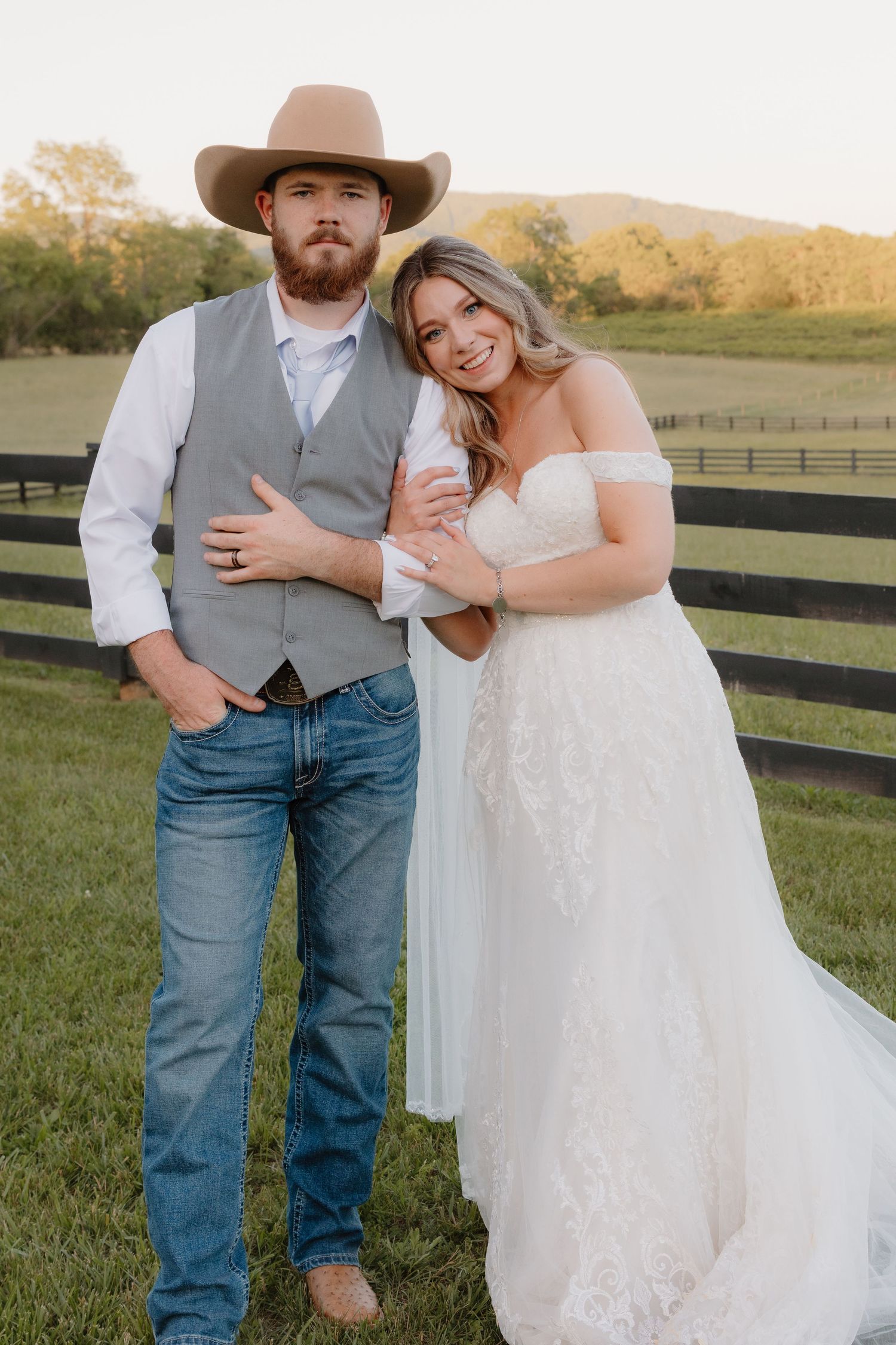 A couple in country wedding attire embraces on a farm with black fences and green grass at sunset.