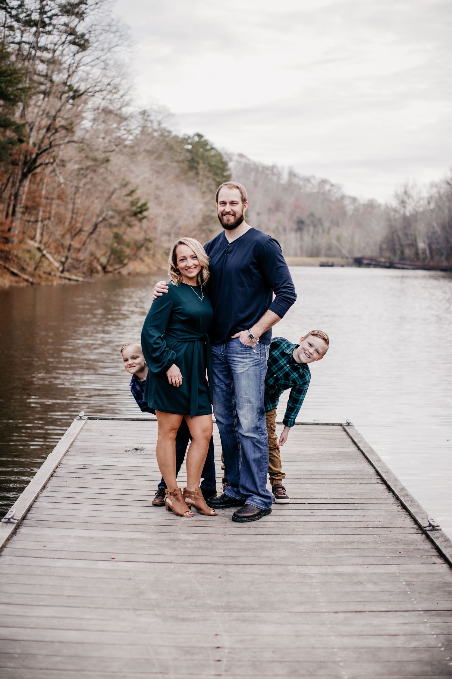A family poses together on a wooden dock overlooking a scenic lake surrounded by winter trees on a cloudy day.