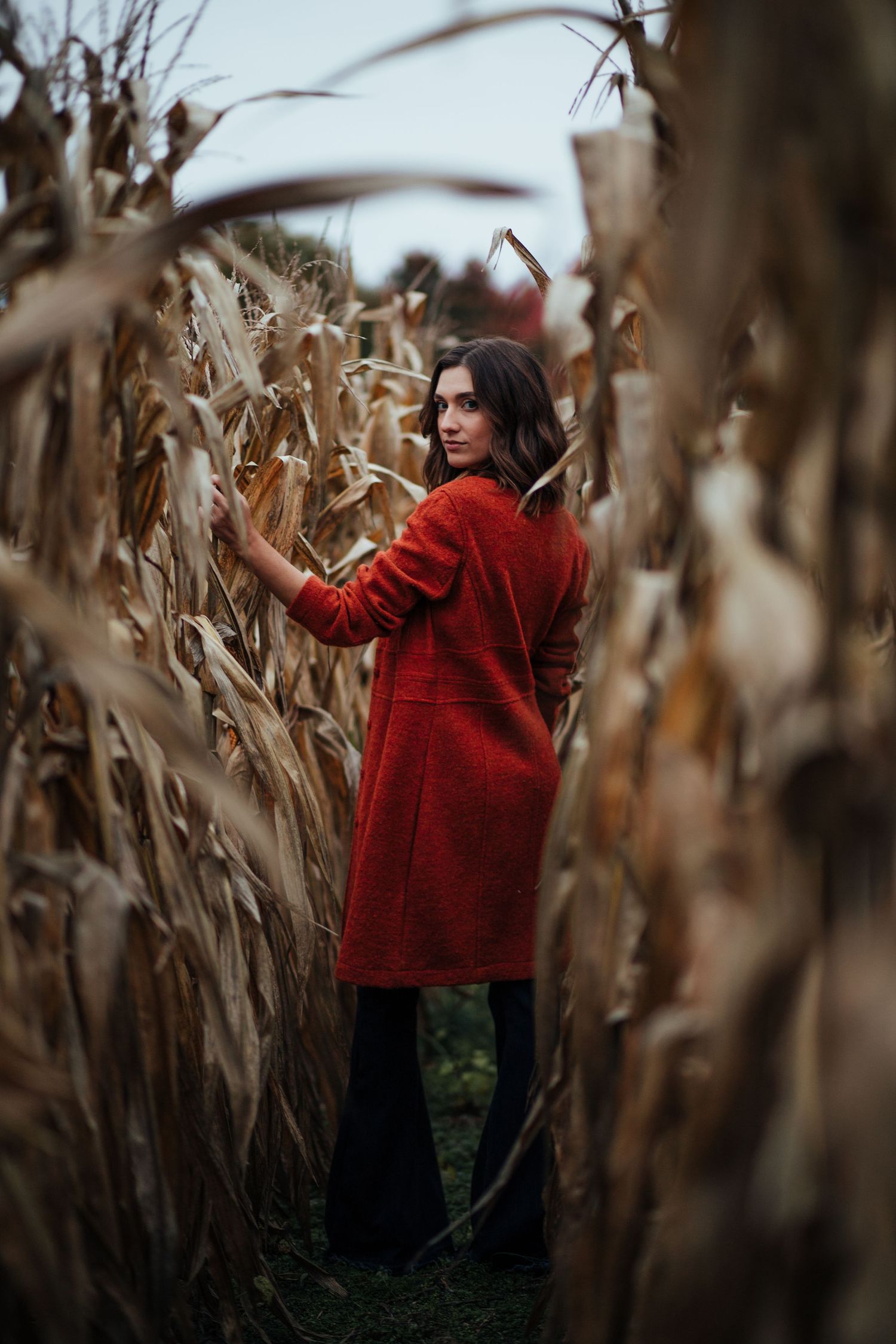 Woman in a striking red coat poses thoughtfully among dried corn stalks.