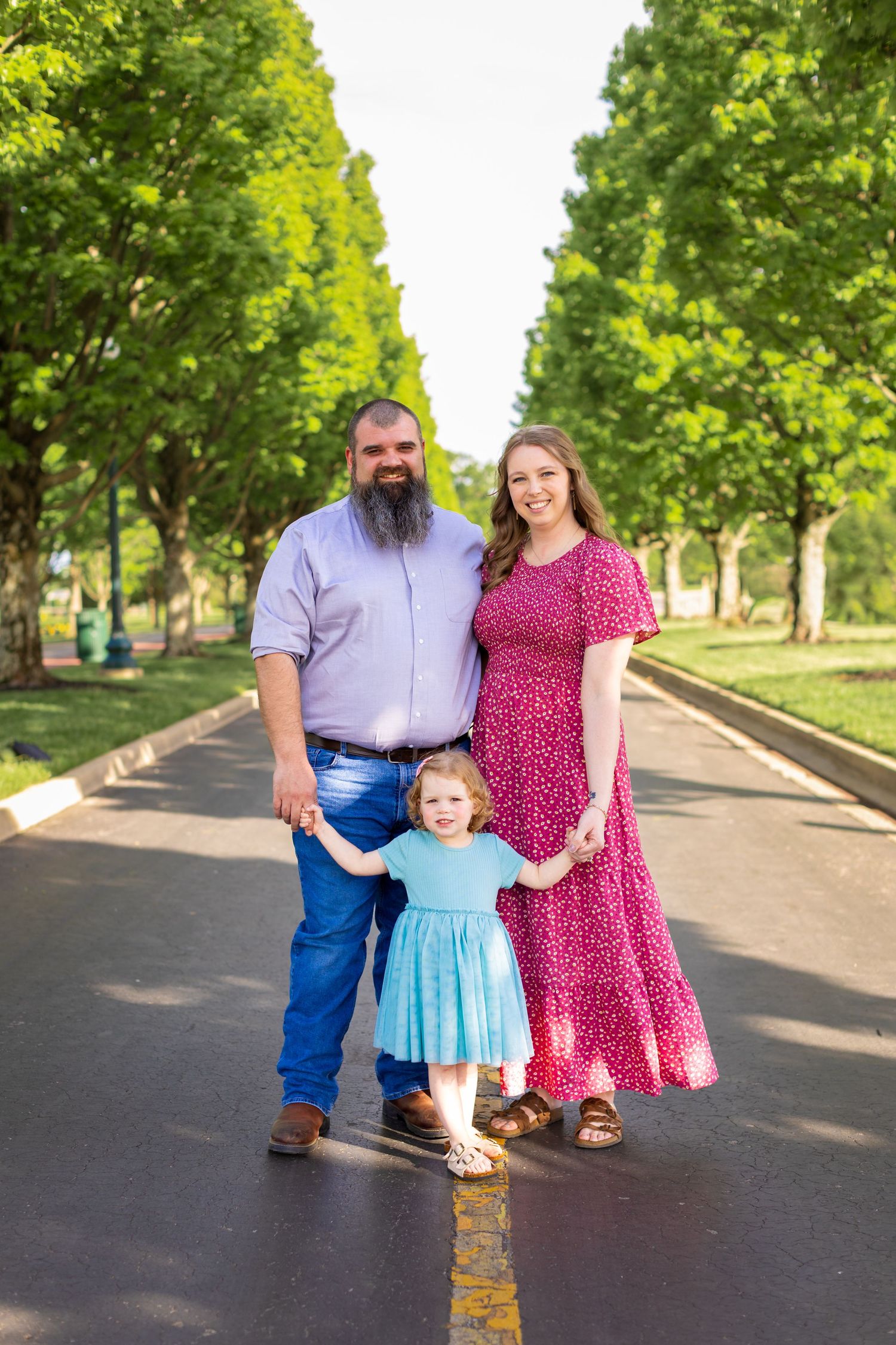Family in coordinating pink and blue outfits takes photos outdoors on a tree-lined path.