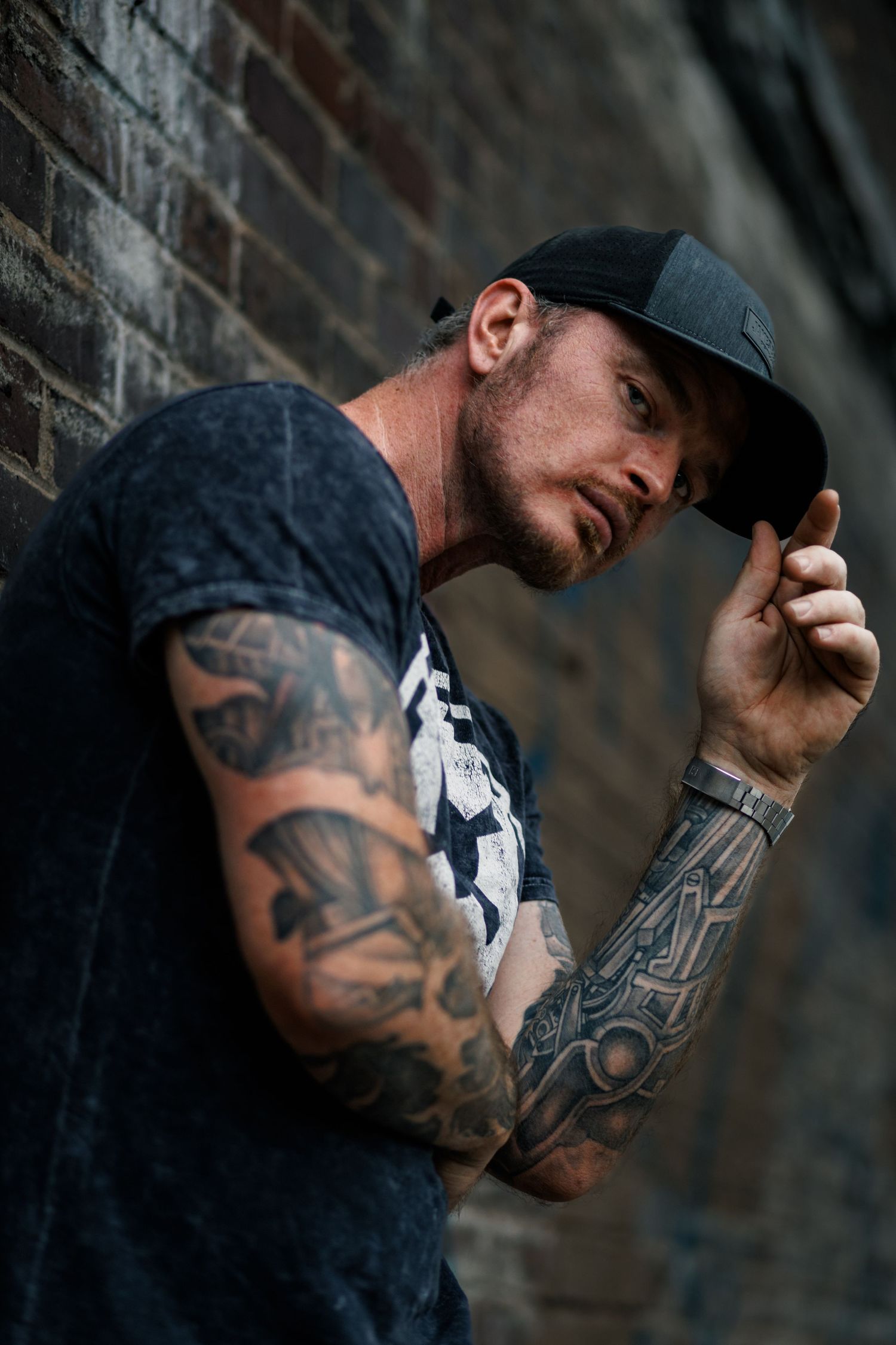 Artistic portrait series showing a person in a dark cap with tattoos against a brick wall in dramatic lighting.