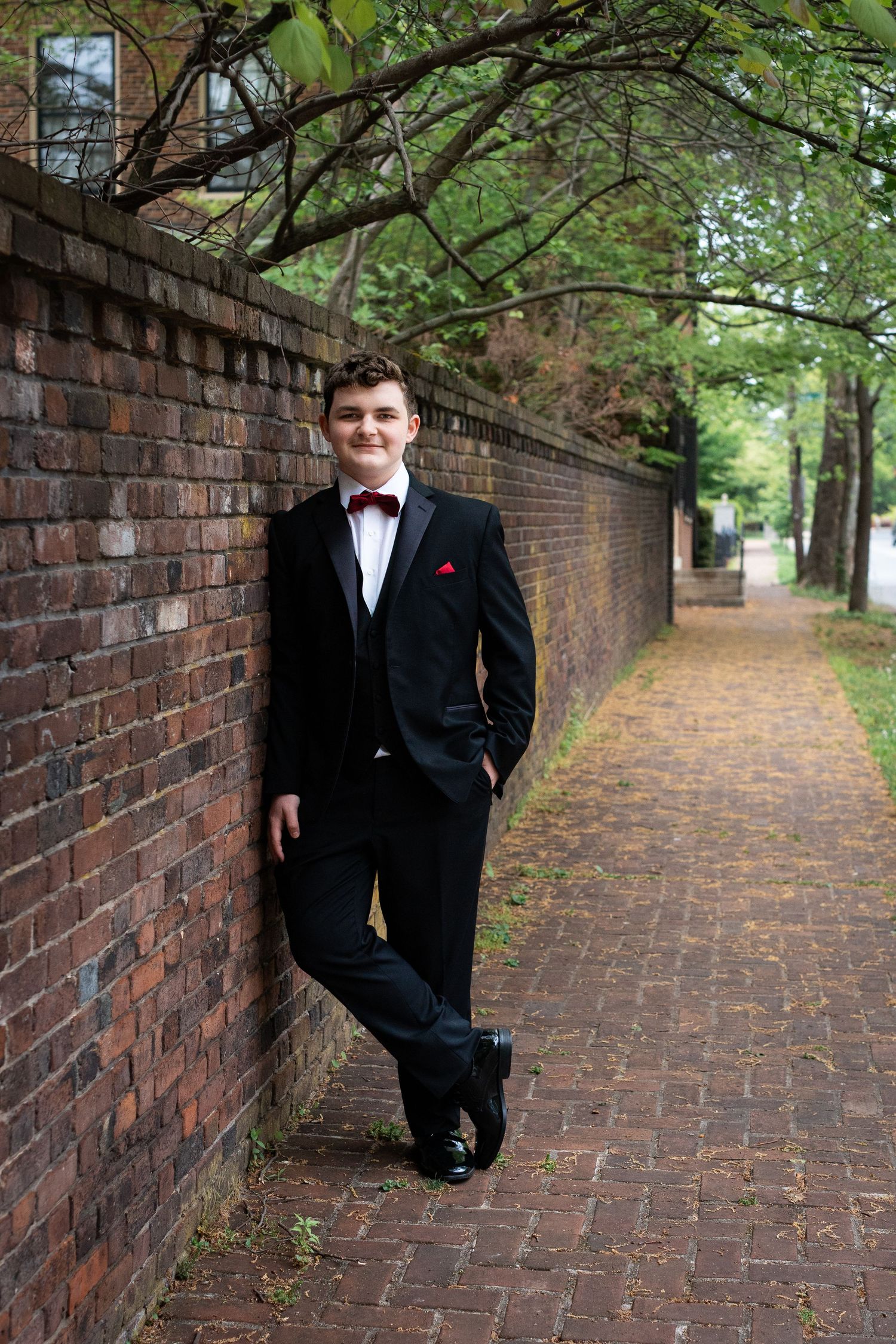 Formal portrait of someone in a black tuxedo and bow tie leaning against a brick wall on a tree-lined path.