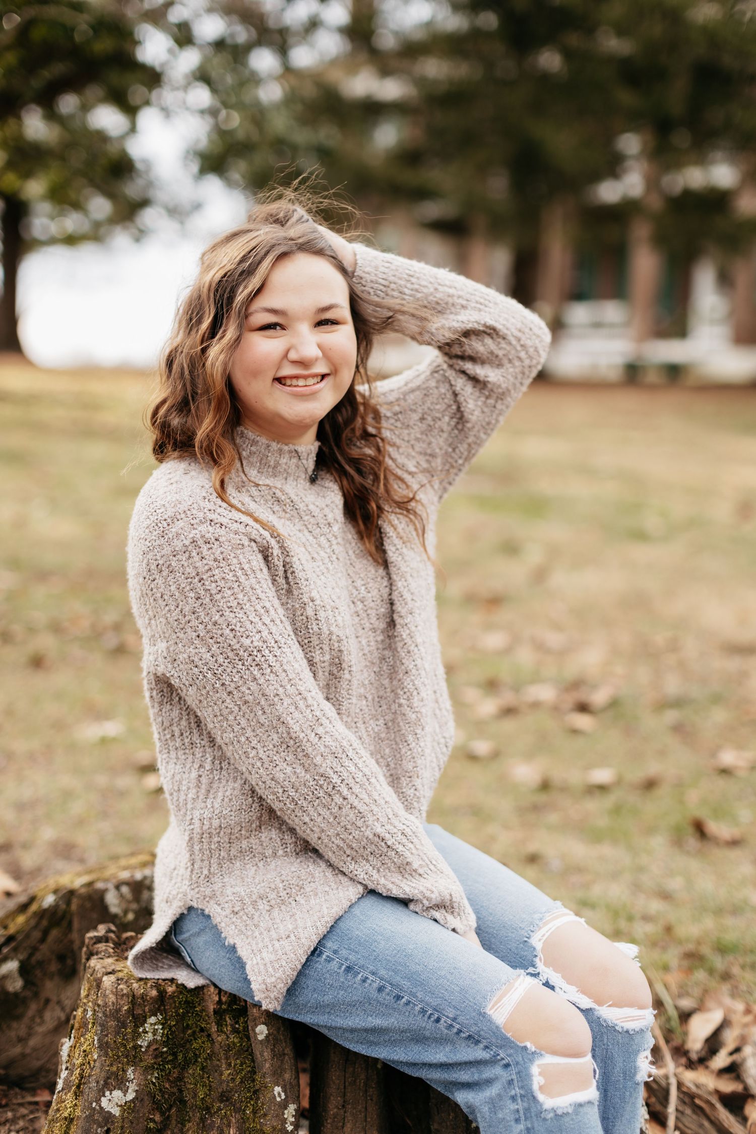 Casual outdoor portrait with someone wearing beige sweater and ripped jeans sitting on tree stump.
