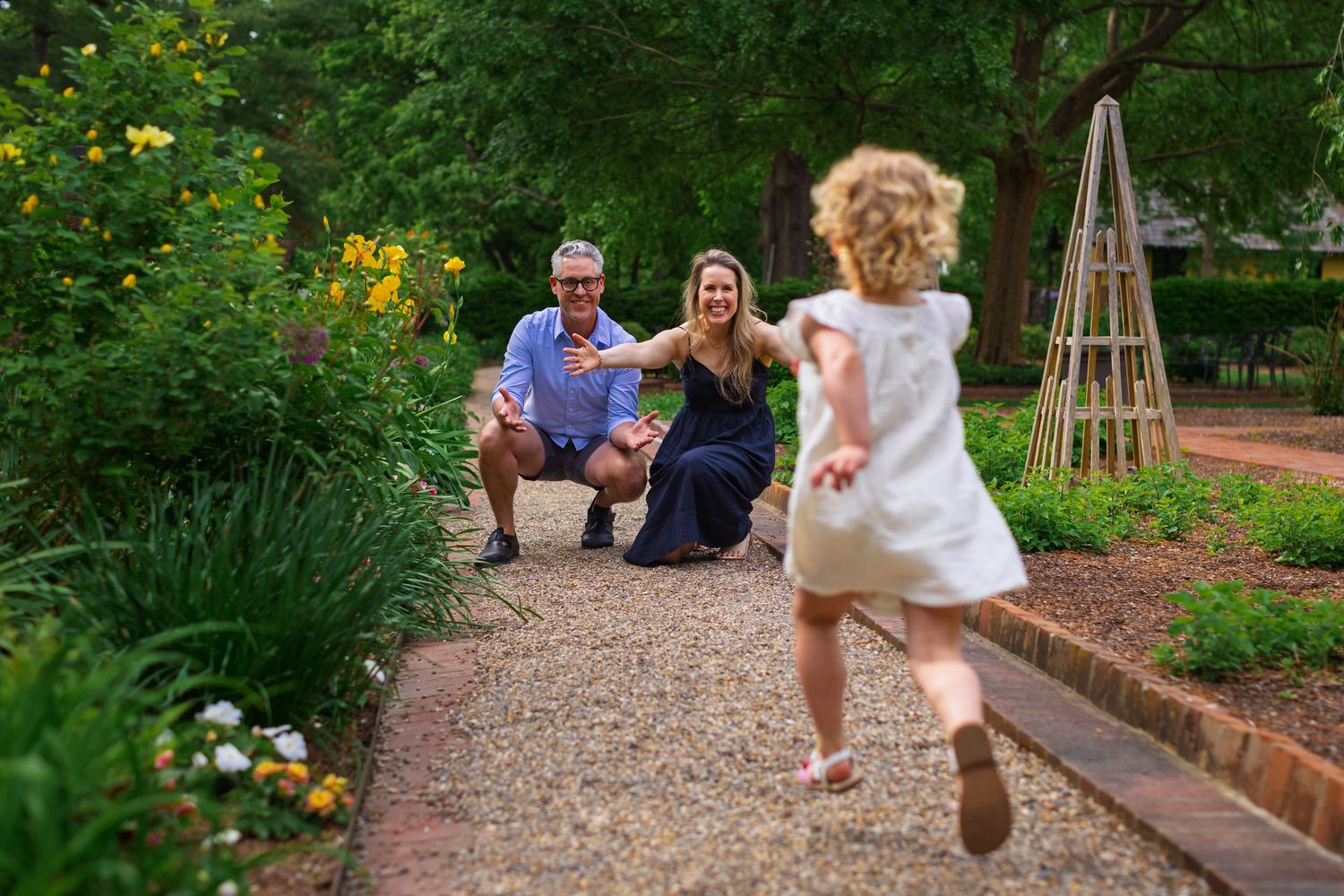 A couple in elegant attire walks together on a gravel garden path surrounded by lush greenery.