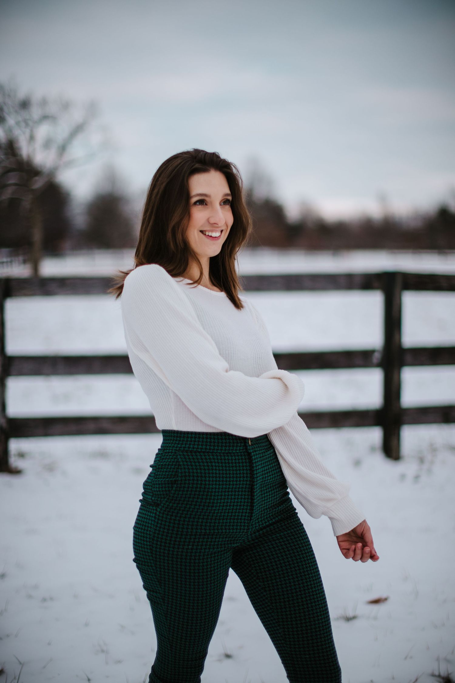 Winter fashion portrait with white sweater and green pants against snowy fence backdrop.